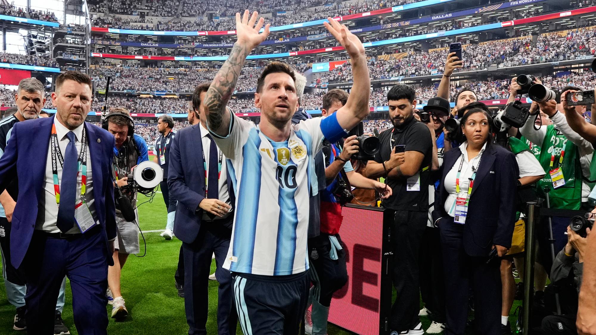 Lionel Messi vinker til publikum på en fotballbane. Han er iført Argentinas drakt og omringes av pressefolk og sikkerhetsvakter. En stor tribunedekket stadion med mange tilskuere er synlig i bakgrunnen. Bildet viser Messi etter en kamp, muligens en seier eller avslutning på et mesterskap. (Bildebeskrivelsen er laget av en KI-tjeneste)