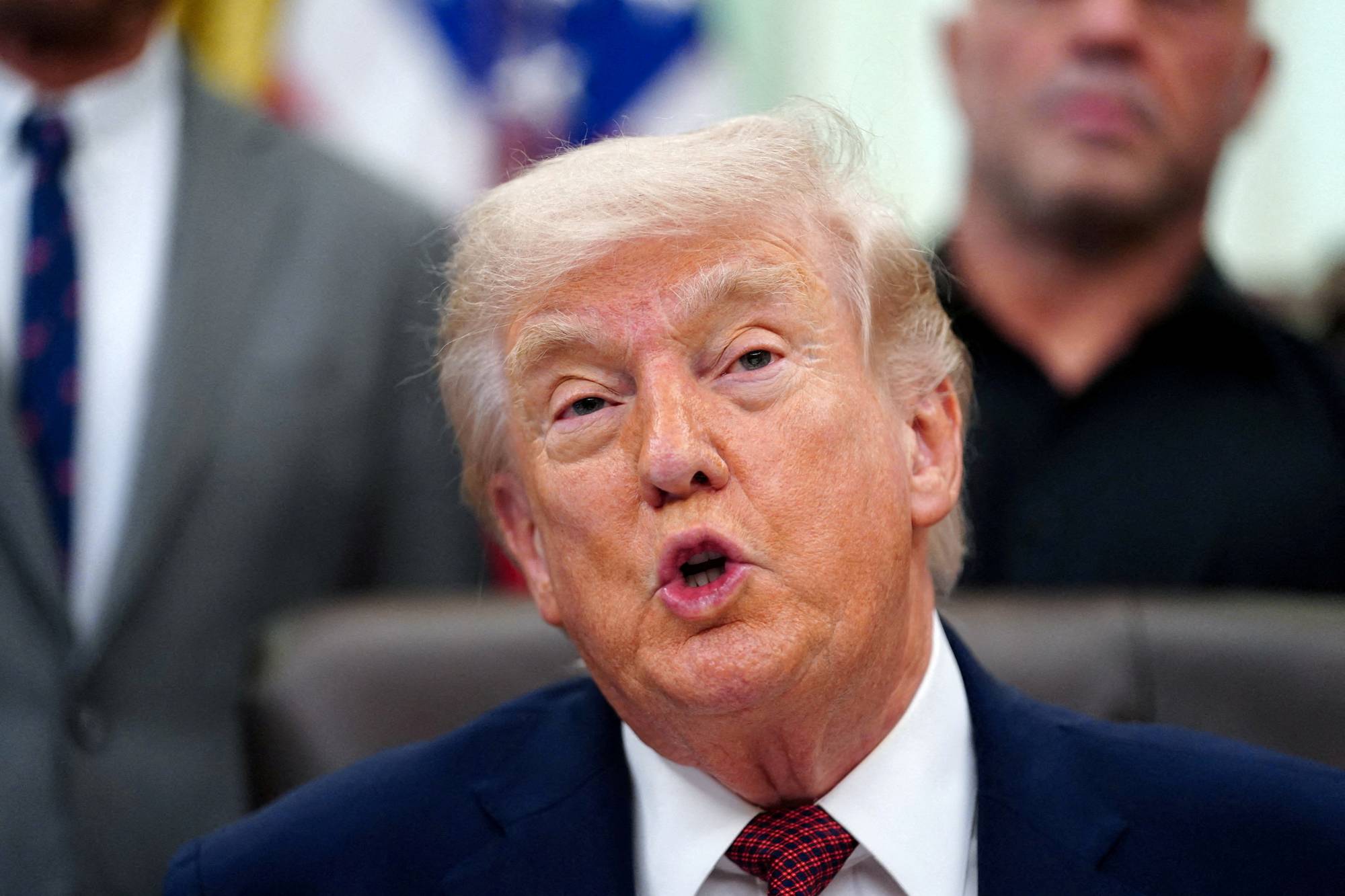 Donald Trump sitter i en stol, med munnen åpen og blikket rettet fremover. En mann i mørk skjorte står i bakgrunnen til høyre. En annen mann i dress og slips er synlig til venstre. (Bildebeskrivelsen er laget av en KI-tjeneste)