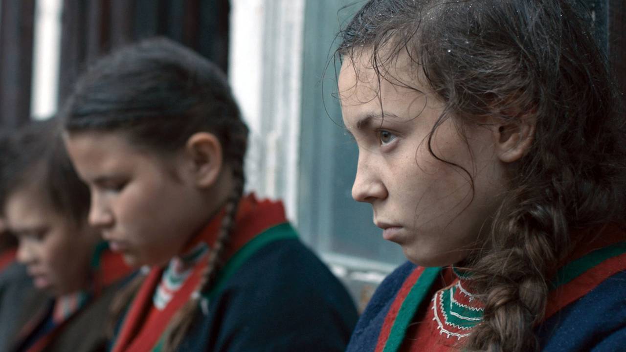 Fra filmen Sameblod. Tre jenter sitter ved siden av hverandre, sett fra siden, jenta foran ser opp på noen, de neste har blikket senket., Iført samiske klær.