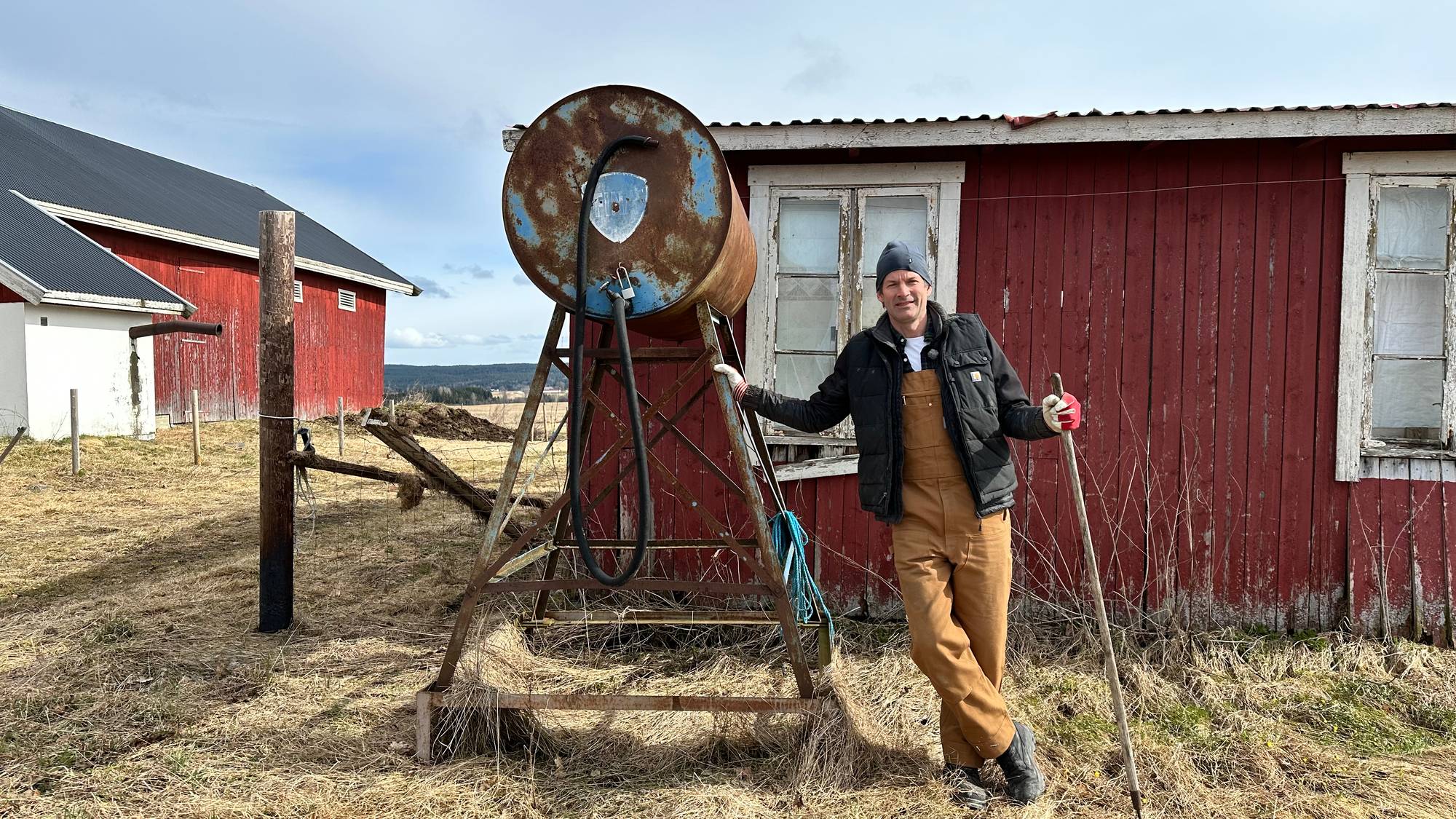 En mann står foran et rødt hus og lener seg inntil en konstruksjon. I hånda holder han en spade.