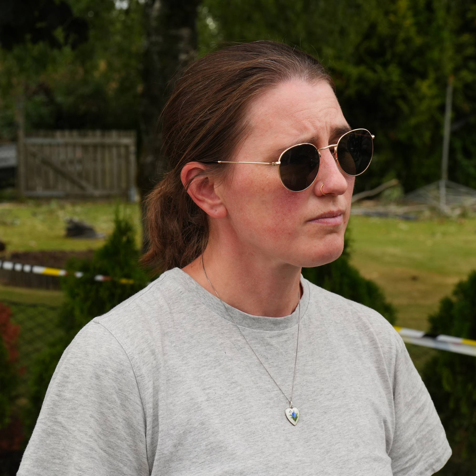 Kvinne med solbriller står utendørs ved avsperret område med skader i bakgrunnen. (Bildebeskrivelsen er laget av en KI-tjeneste)