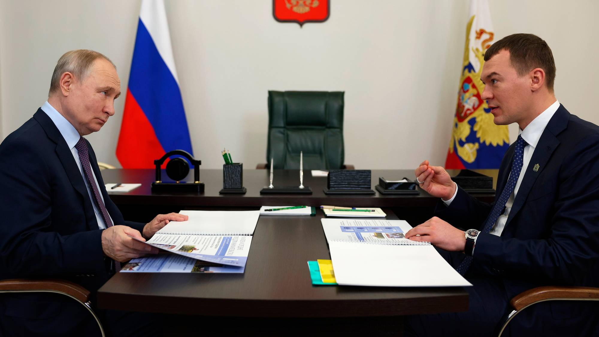 Vladimir Putin sitter ved et skrivebord og samtaler med Mikhail Degtjarjov foran russiske flagg. (Bildebeskrivelsen er laget av en KI-tjeneste)