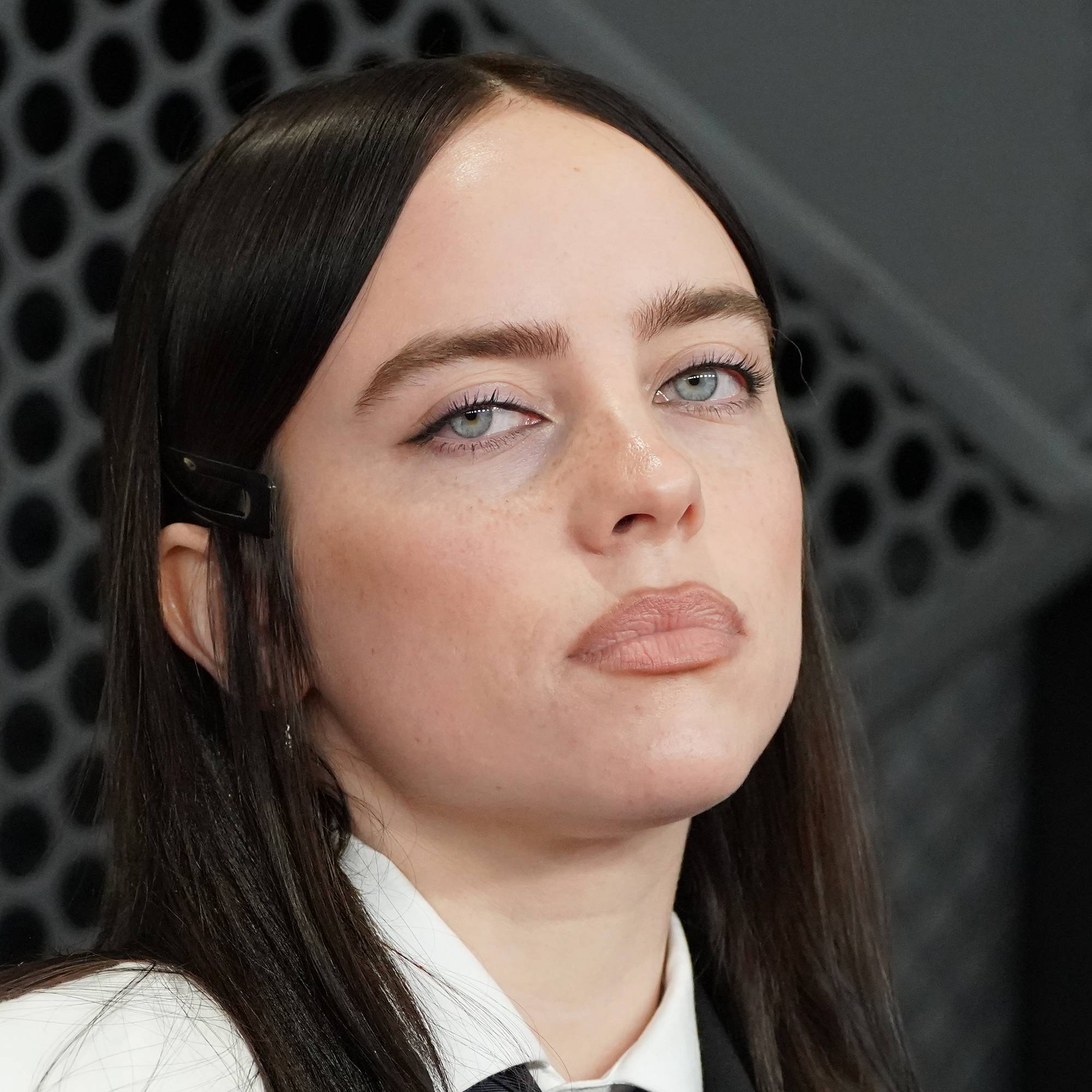 Amerikanske Billie Eilish deltar på en begivenhet. Hun har mørkt hår, med en hårspenne i venstre side. Sangeren har på seg hvit skjorte med mørkeblå slips. Hun har en svart vest over skjorta. Bak henne er en grå bakgrunn med et mønster av runde hull. (Bildebeskrivelsen er laget av en KI-tjeneste)