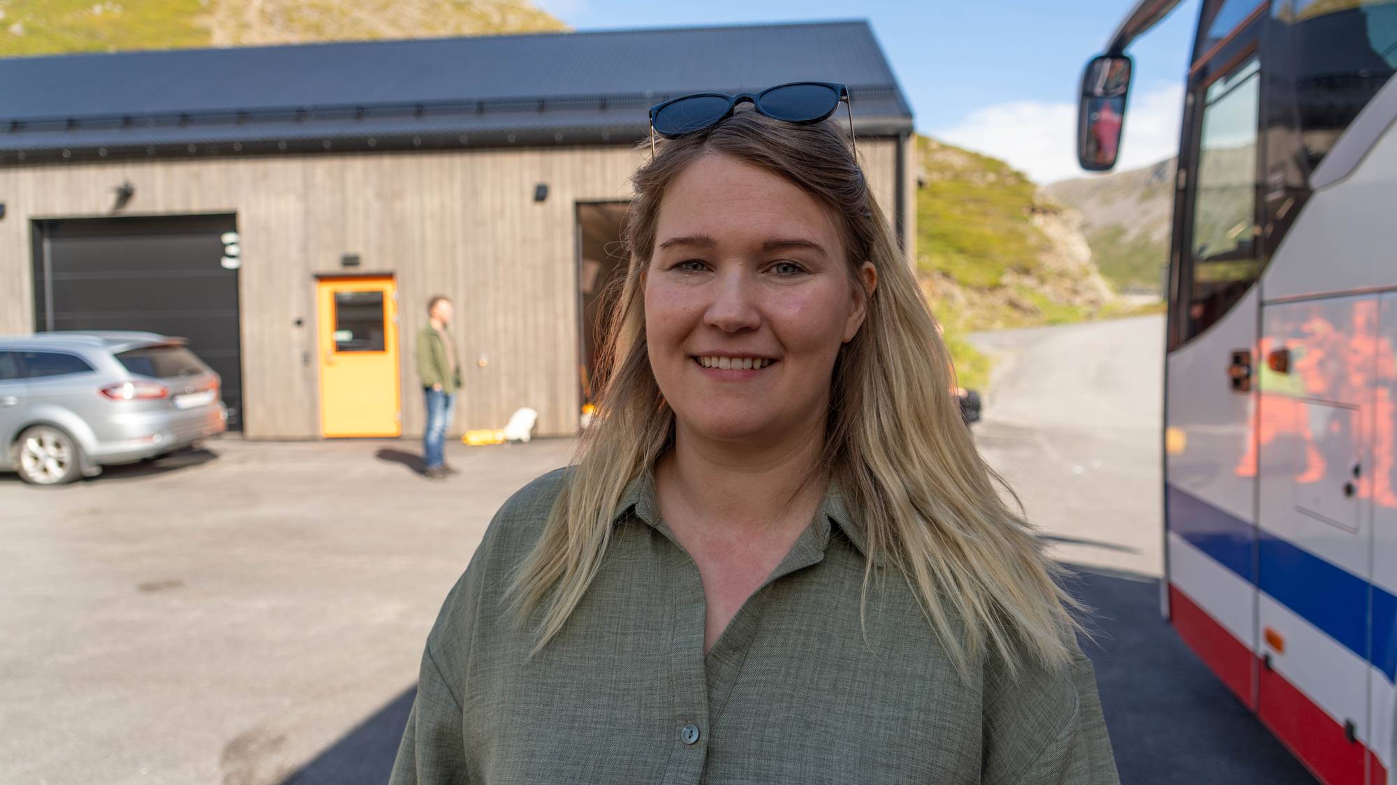 Kvinne med solbriller på hodet smiler. En bygning og en buss er synlige i bakgrunnen. (Bildebeskrivelsen er laget av en KI-tjeneste)