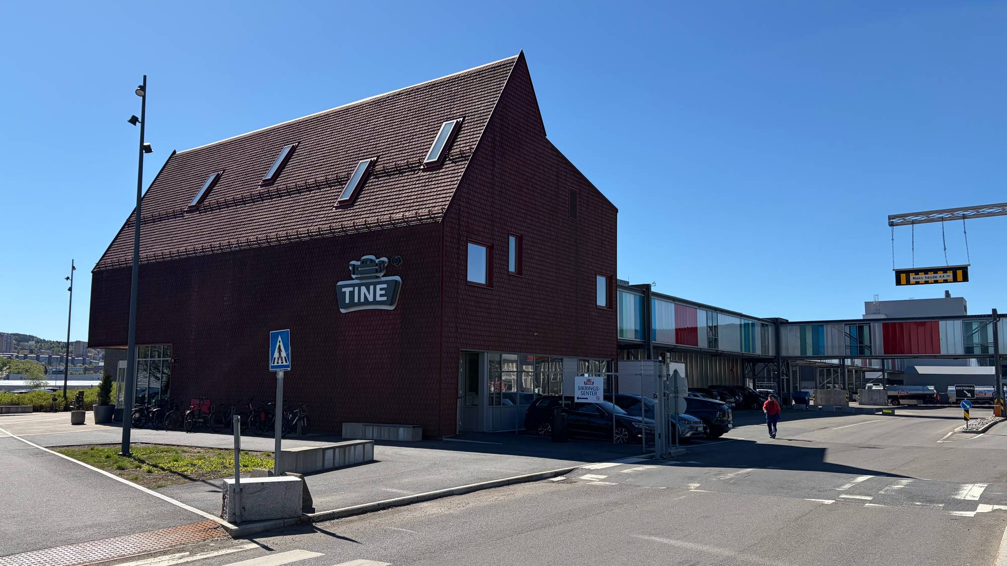 Bygning med Tine-logo. En person går mot en tilknyttet bygningsdel. Flere biler er parkert utenfor. Det er en krysning i forgrunnen. (Bildebeskrivelsen er laget av en KI-tjeneste)
