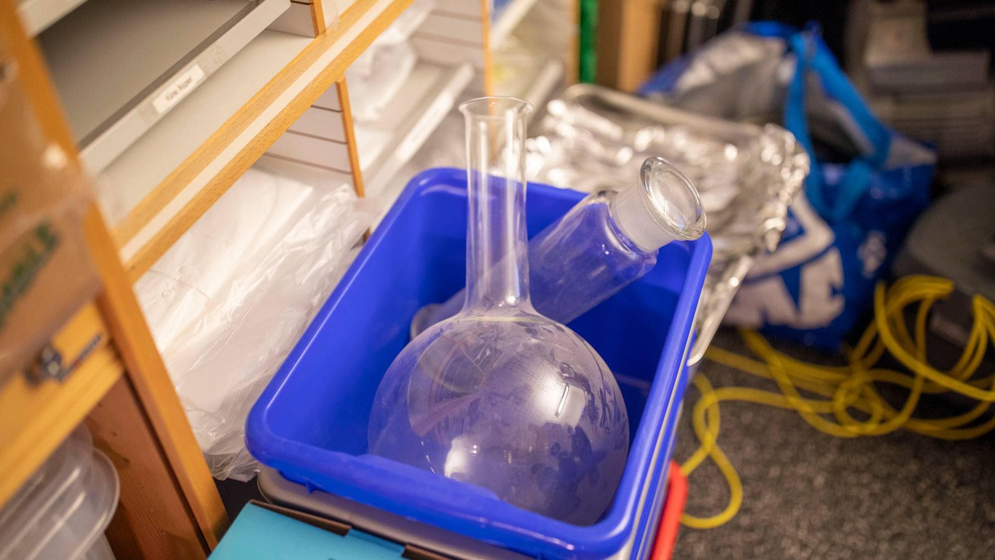 En blå plastkasse inneholder to runde glasskolber. Kolbene er tomme og har korker i halsen. Kassen står i et rom med hyller, poser og ledninger i bakgrunnen. (Bildebeskrivelsen er laget av en KI-tjeneste)