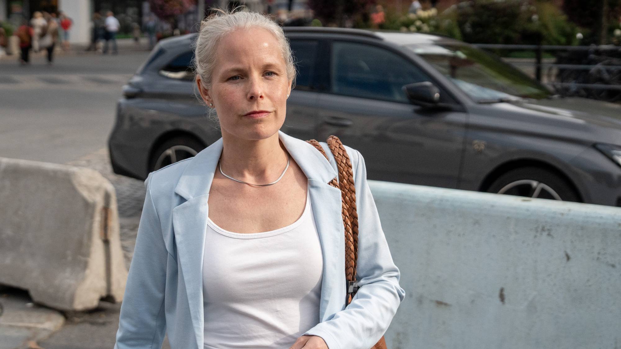 Kvinne med lyseblå blazer og hvit topp går mot kamera. Over venstre skulder henger en brun skulderveske.
