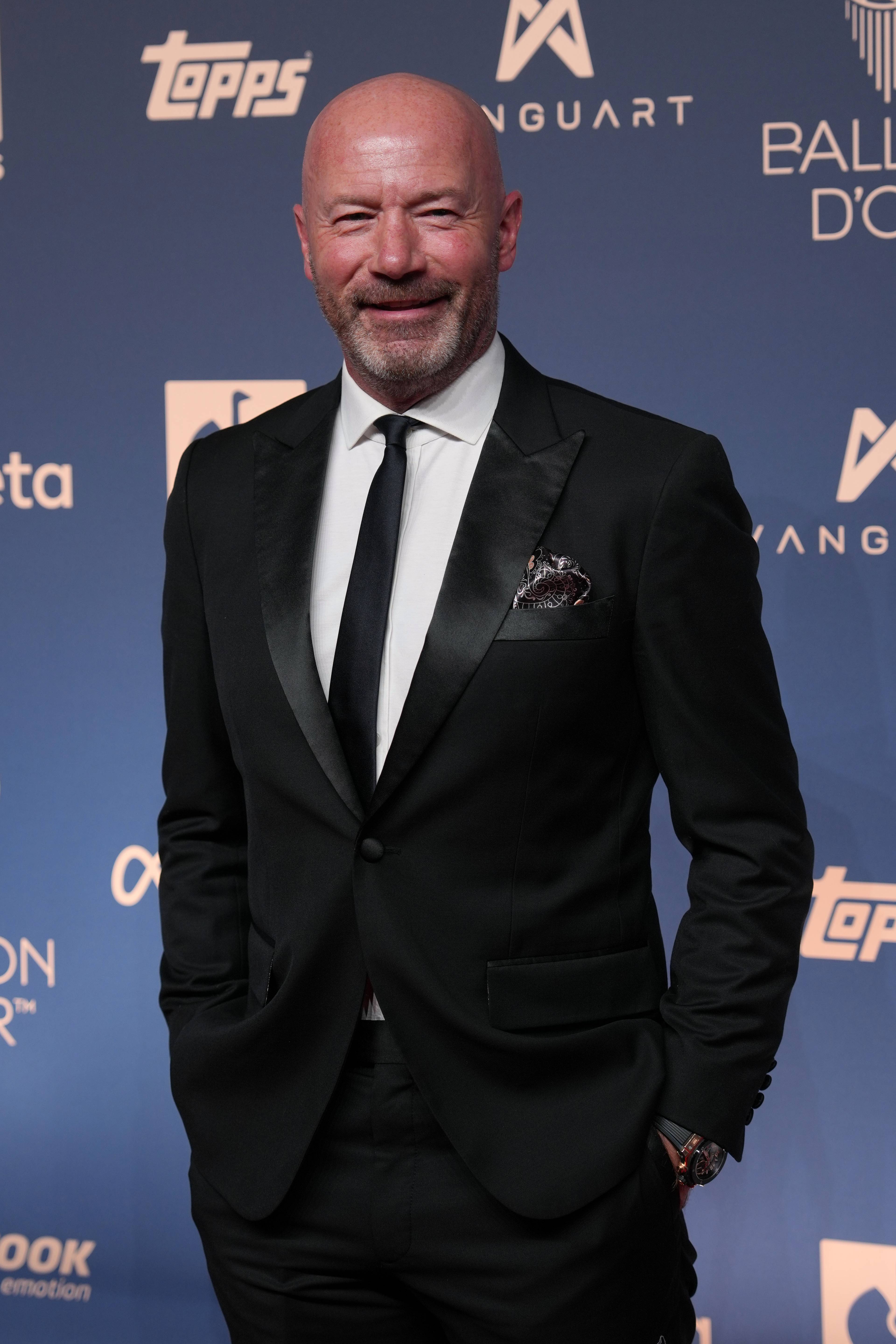 Alan Shearer i dress og slips foran en blå fotovegg med logoer. (Bildebeskrivelsen er laget av en KI-tjeneste)