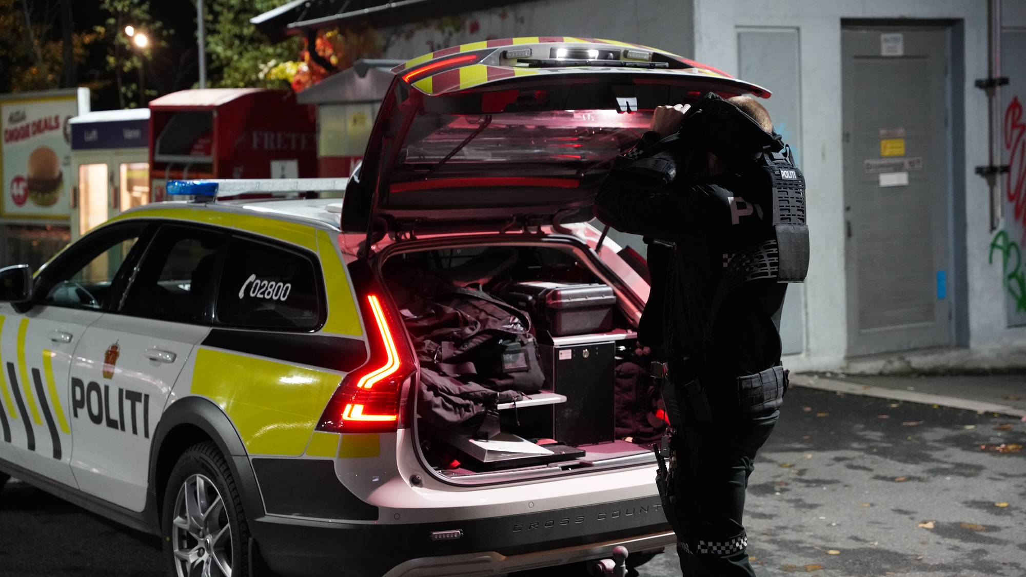 Politibetjent tar på seg utstyr ved en politibil med åpent bagasjerom om kvelden. (Bildebeskrivelsen er laget av en KI-tjeneste)