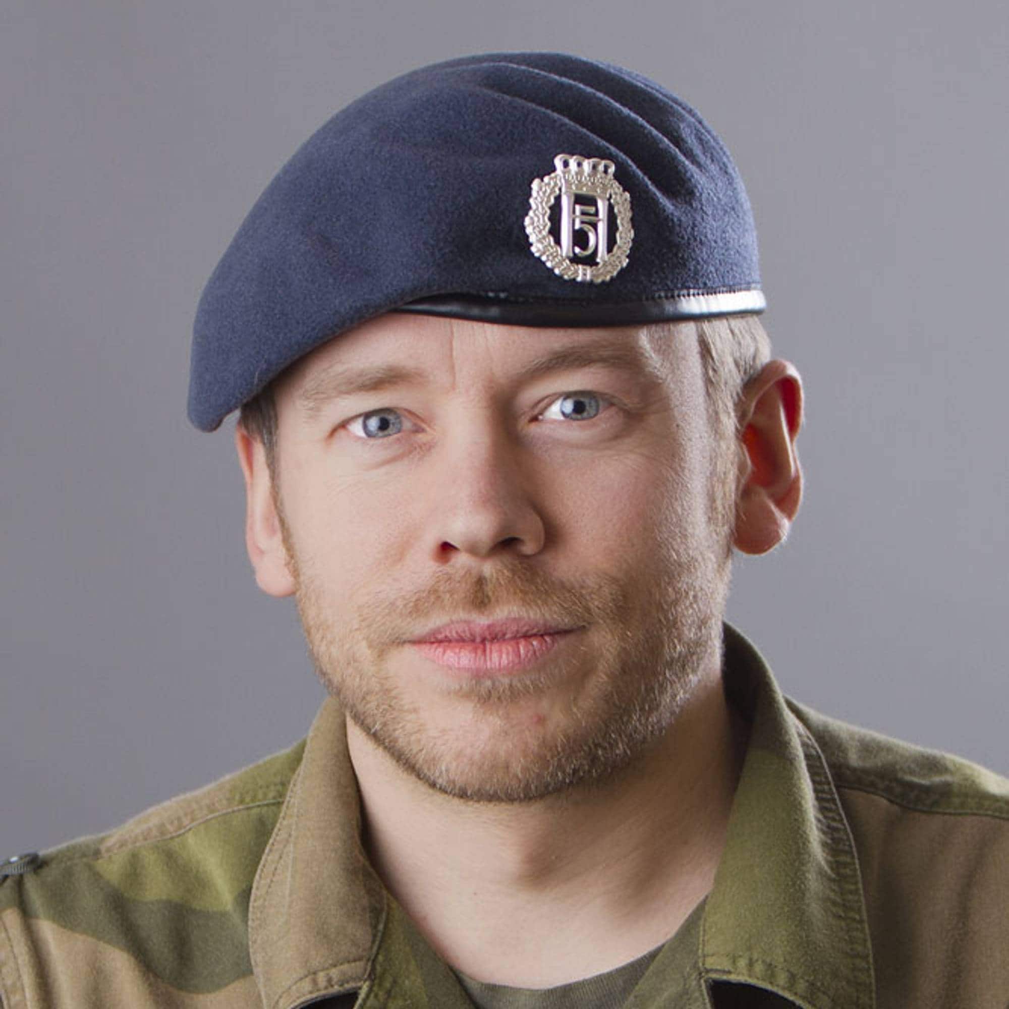 Et portrett av en mann med blå beret og kamuflasjeuniform. Mannen har blå øyne og skjeggstubb. På uniformen er det et navneskilt med teksten STORDAL. (Bildebeskrivelsen er laget av en KI-tjeneste)