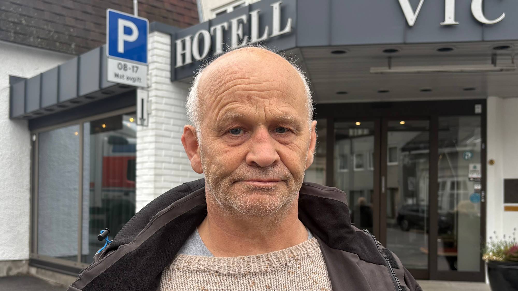 En mann med kort, hvitt hår på sidene, lys genser og brun jakke står utenfor et hotell.