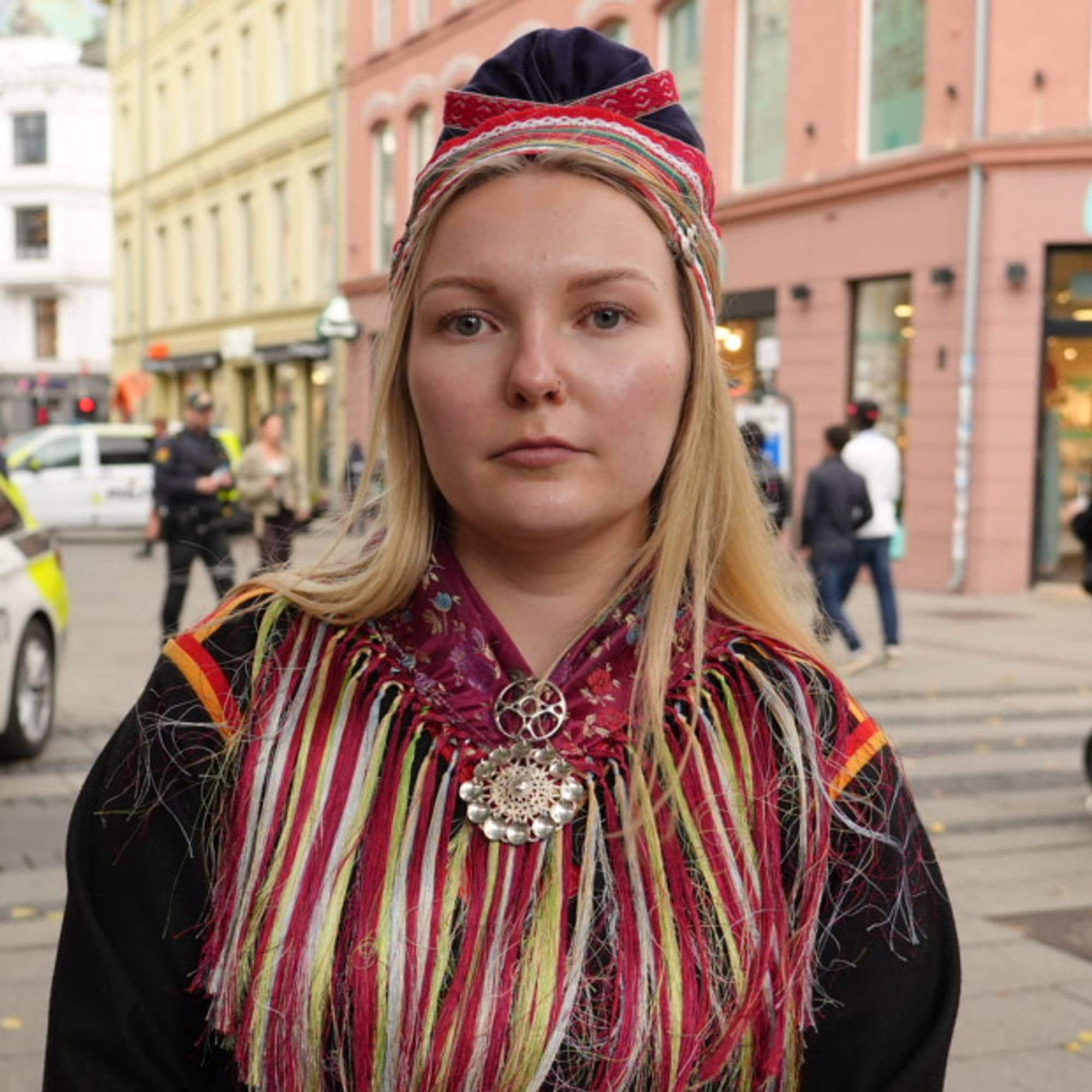 Kvinne i samisk kofte foran en politibil i en bygate med fotgjengere. (Bildebeskrivelsen er laget av en KI-tjeneste)