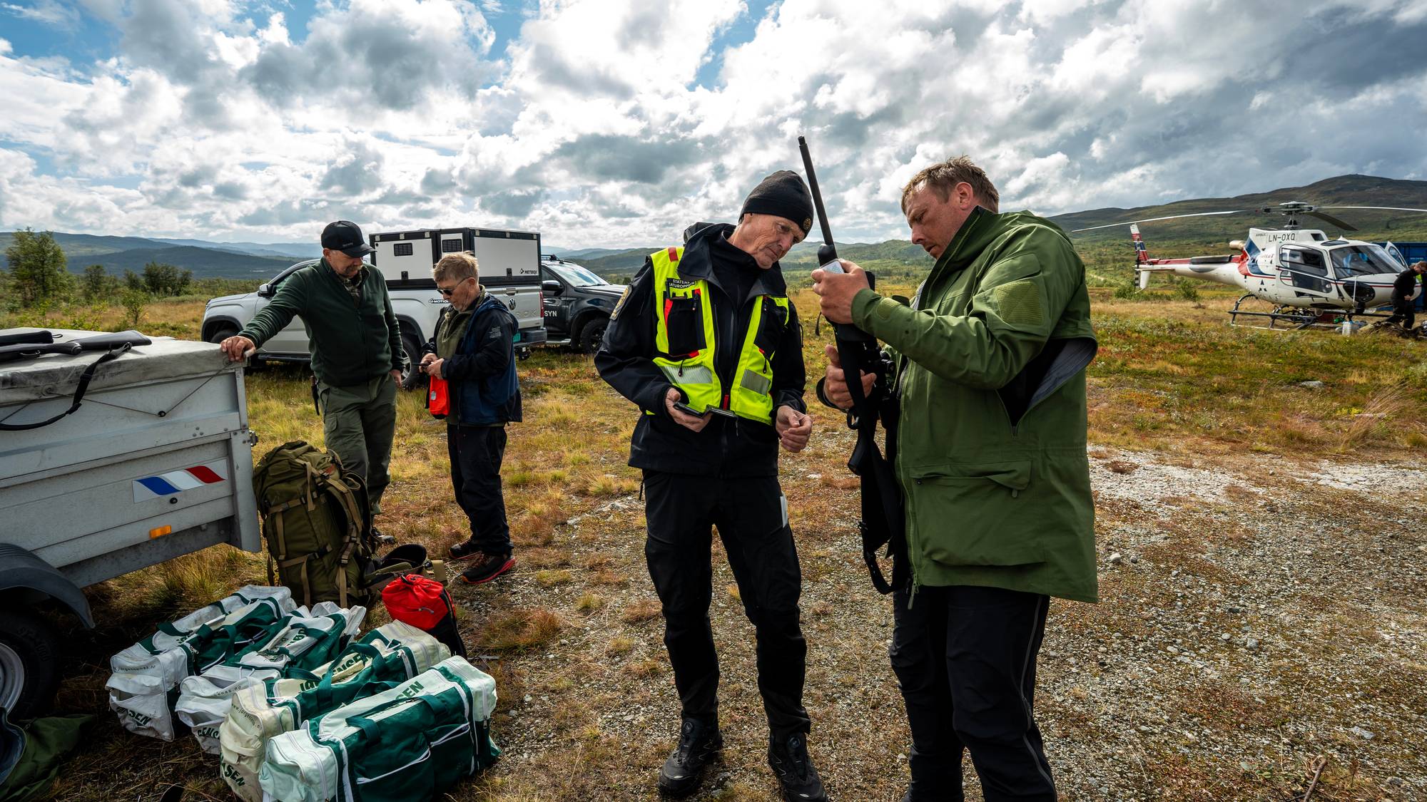 Menn med utstyr ved et helikopter i et fjellandskap. En person kontrollerer et våpen. (Bildebeskrivelsen er laget av en KI-tjeneste)