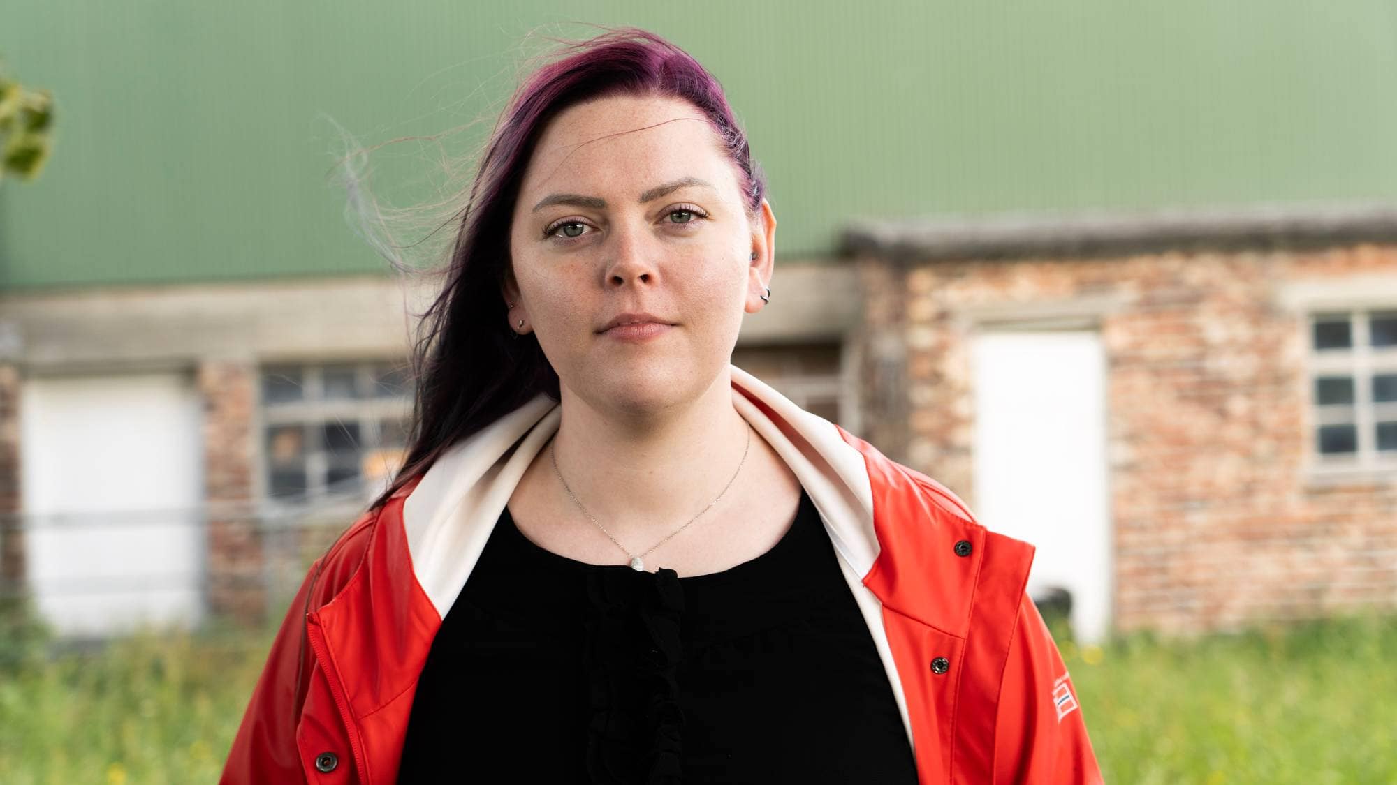 Et bilde av en kvinne med lilla hår, foran en mursteinsbygning. Hun har på seg en rød jakke. (Bildebeskrivelsen er laget av en KI-tjeneste)