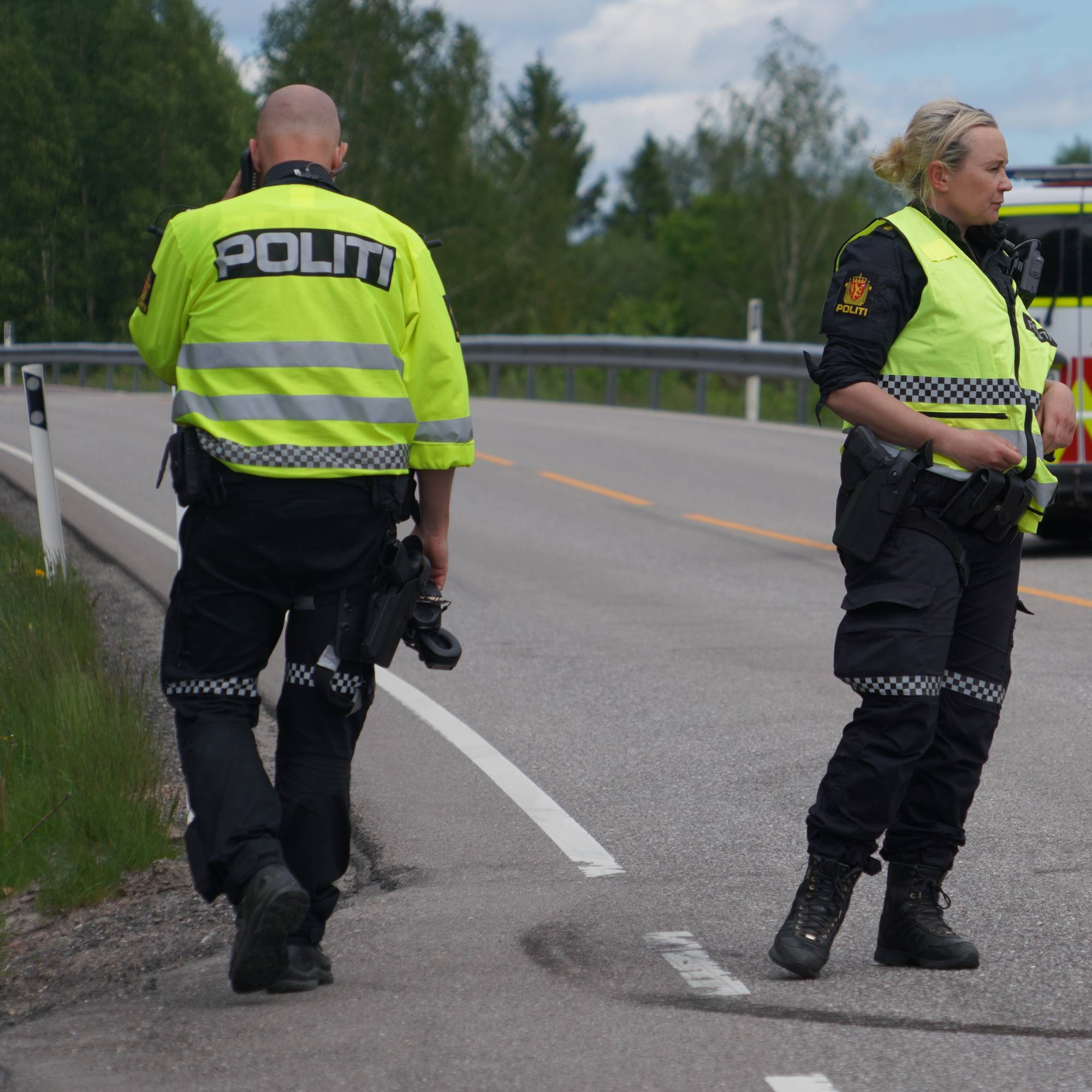 To politibetjenter står ved en landevei, der en politibil står parkert med åpen dør. 