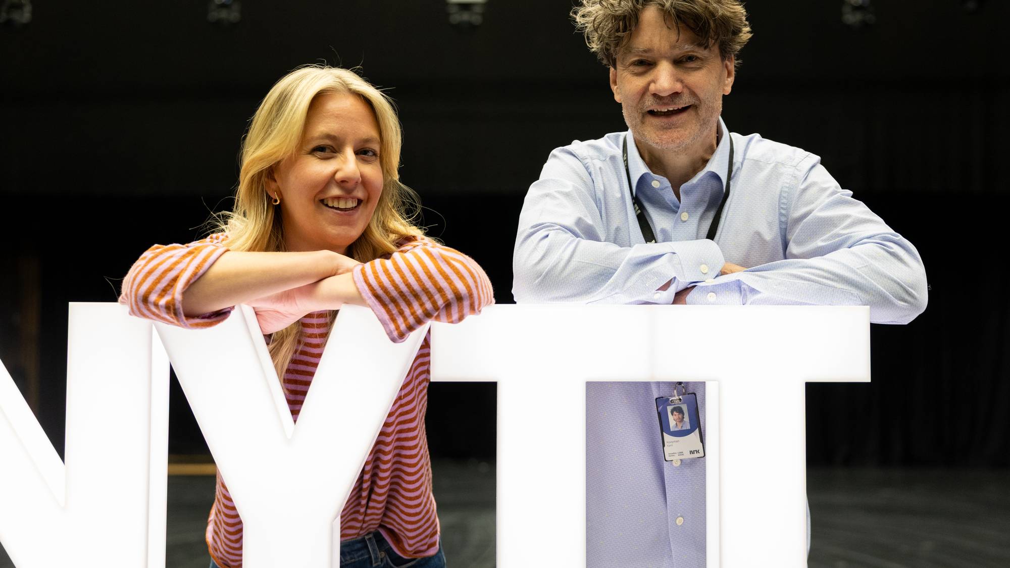 Kristine Grændsen og Kjetil Kooymann står bak et stort, opplyst "NYTT på Nytt"-skilt. Begge smiler mot kamera. Bak dem skimtes et mørkt rom med lysrigg i taket. 