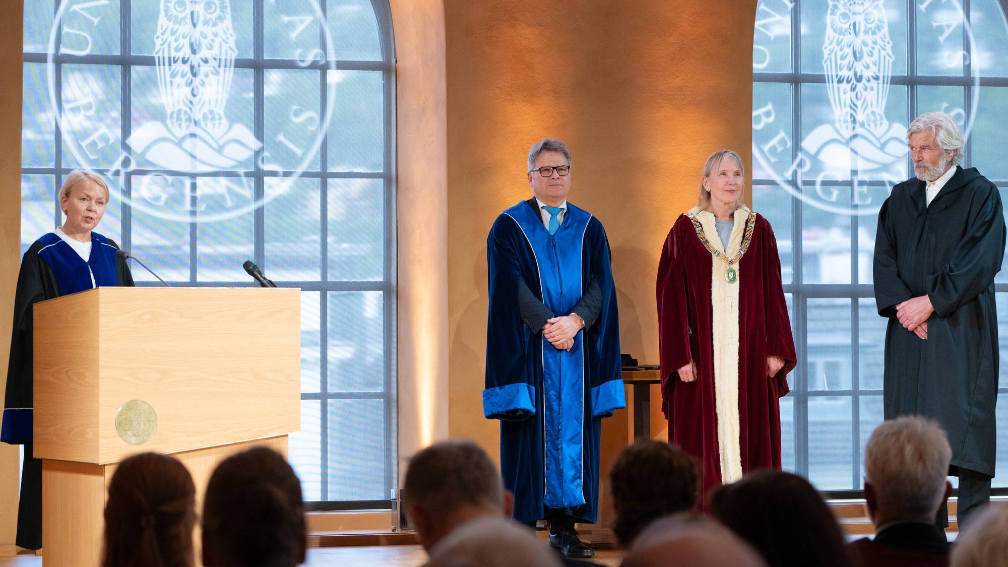dame i blå kappe bak talarstol, litt bortanfor står mann i kappe, dame med rektorkjede og kappe ved sidan av mann i mørk kappe