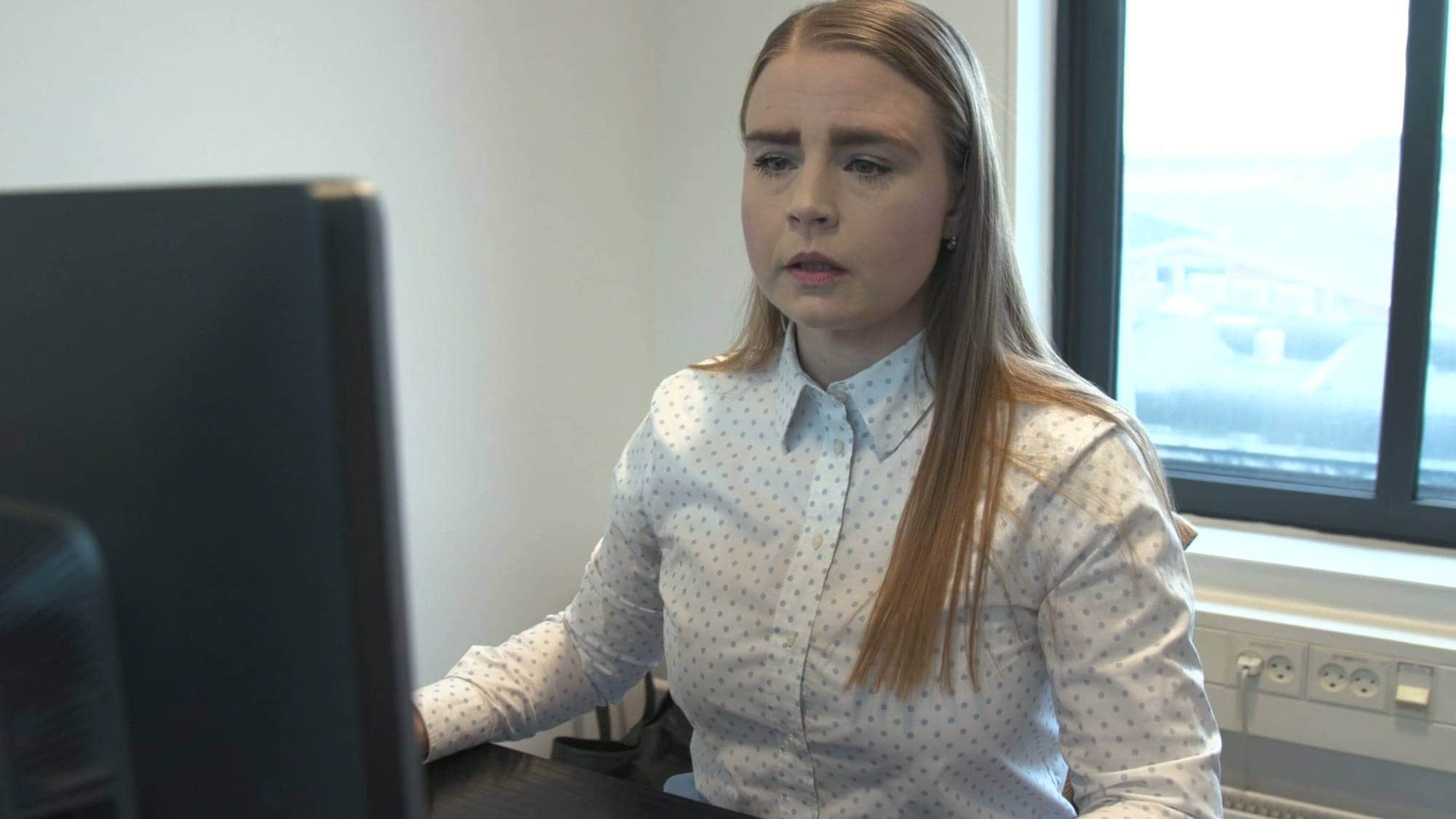 En kvinne med lyst hår sitter ved et skrivebord og ser inn i en dataskjerm. Hun har på seg en hvit skjorte med blå prikker. Bak henne er et vindu med utsikt og stikkontakter på veggen. (Bildebeskrivelsen er laget av en KI-tjeneste)