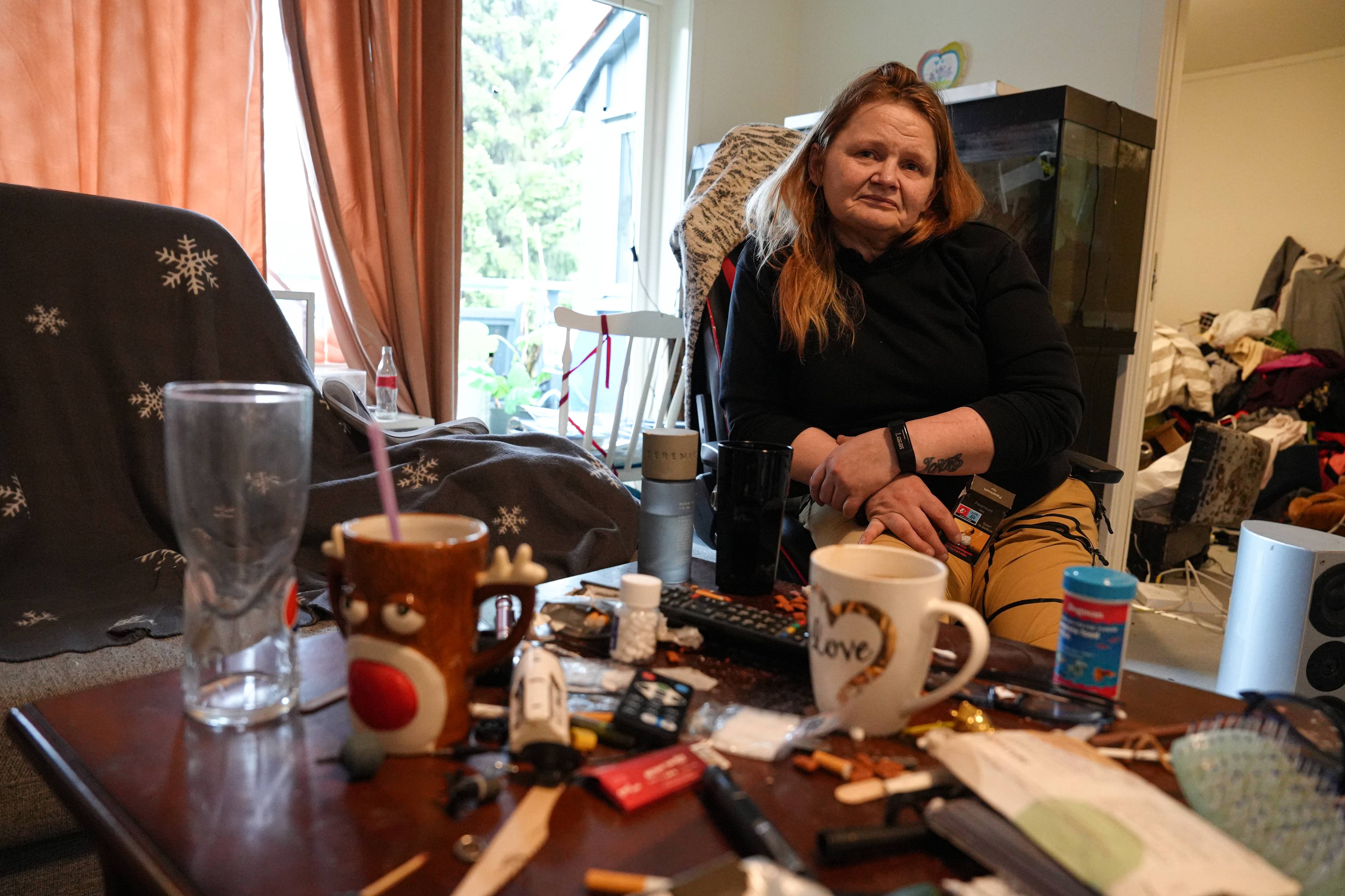 Kvinne sitter på en stol i en stue. Rundt henne er det rot alle steder.