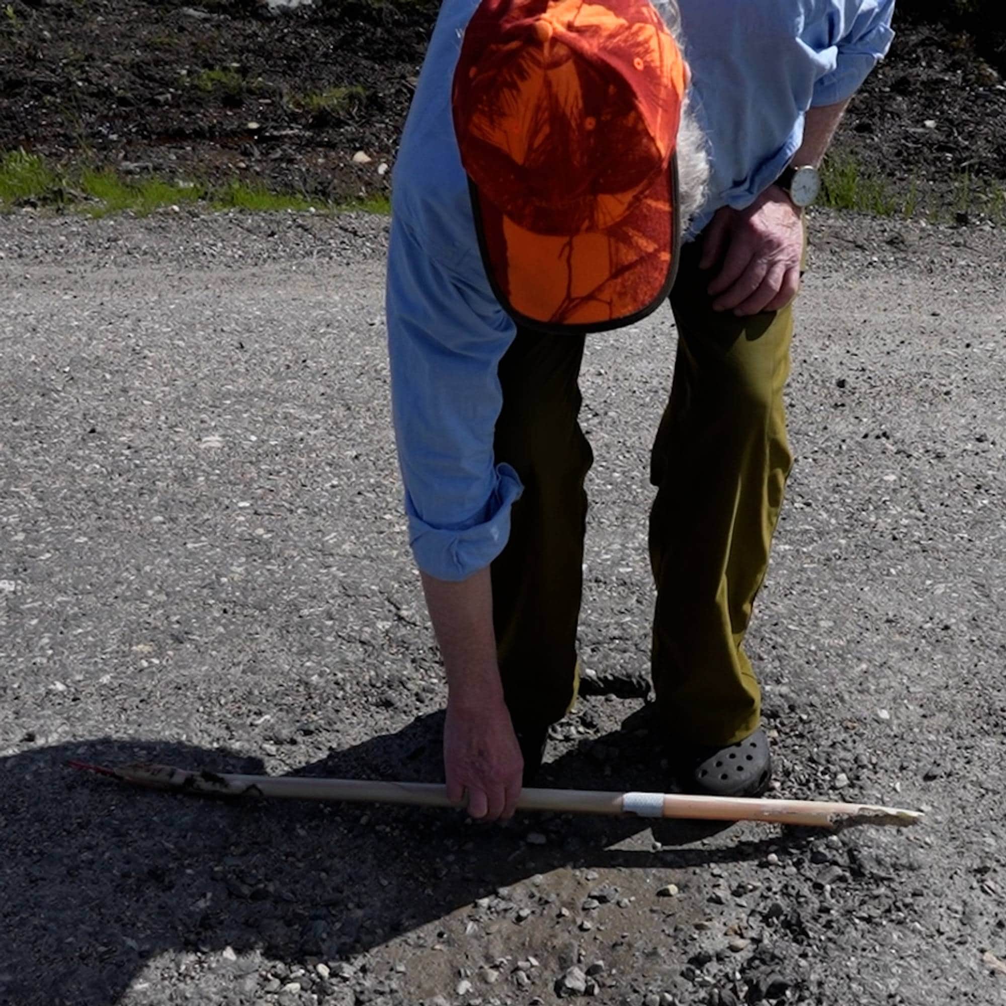 Person i oransje caps måler et hull i en grusvei med en pinne. (Bildebeskrivelsen er laget av en KI-tjeneste)