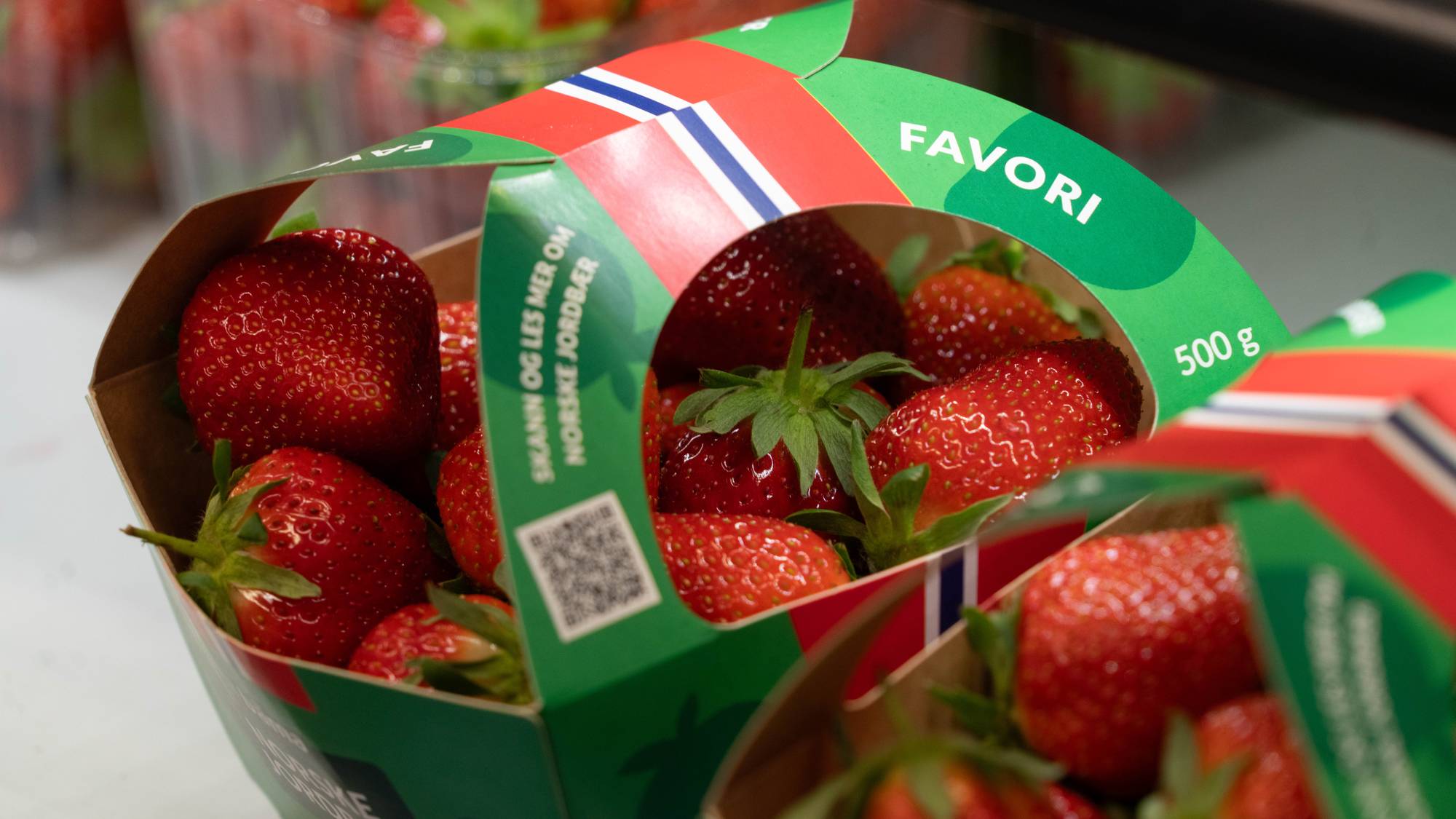 Et parti norske jordbær i pappemballasje er vist. Emballasjen har et grønt bånd med teksten "FAVORI" og "500 g". Den har også et norsk flagg og teksten "SKANN OG LES MER OM NORSKE JORDBÆR". En prisetikett er synlig i bakgrunnen. (Bildebeskrivelsen er laget av en KI-tjeneste)