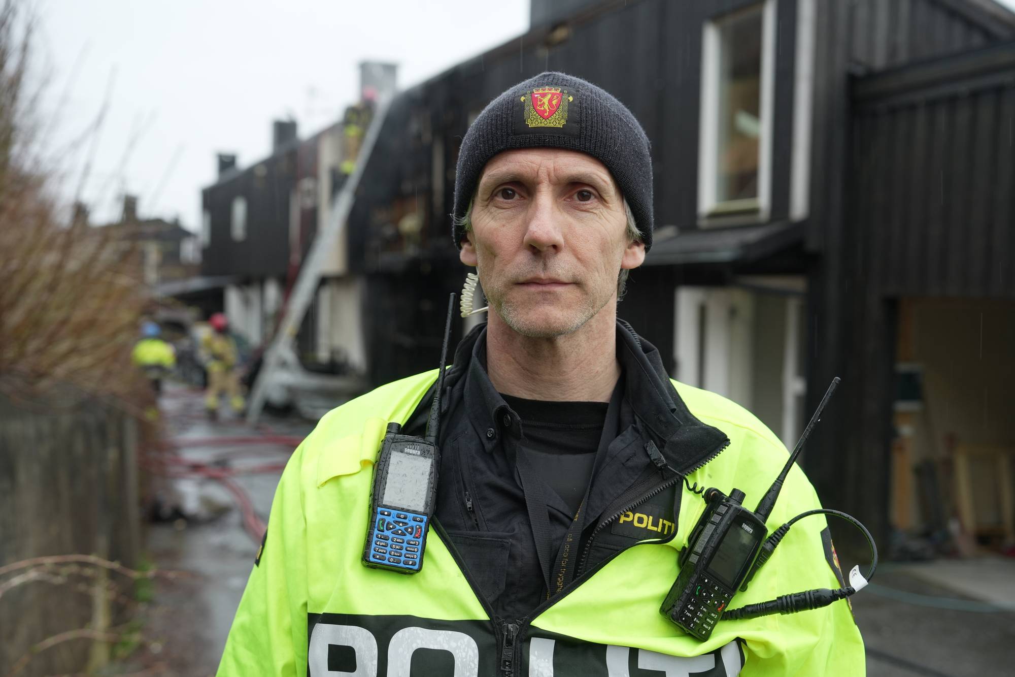 Politimann står foran en nedbrent bygning.
Han har på seg en gul refleksvest og en svart lue.
To politiradioer er festet til vesten.
I bakgrunnen er det andre politifolk og brannslanger. (Bildebeskrivelsen er laget av en KI-tjeneste)
