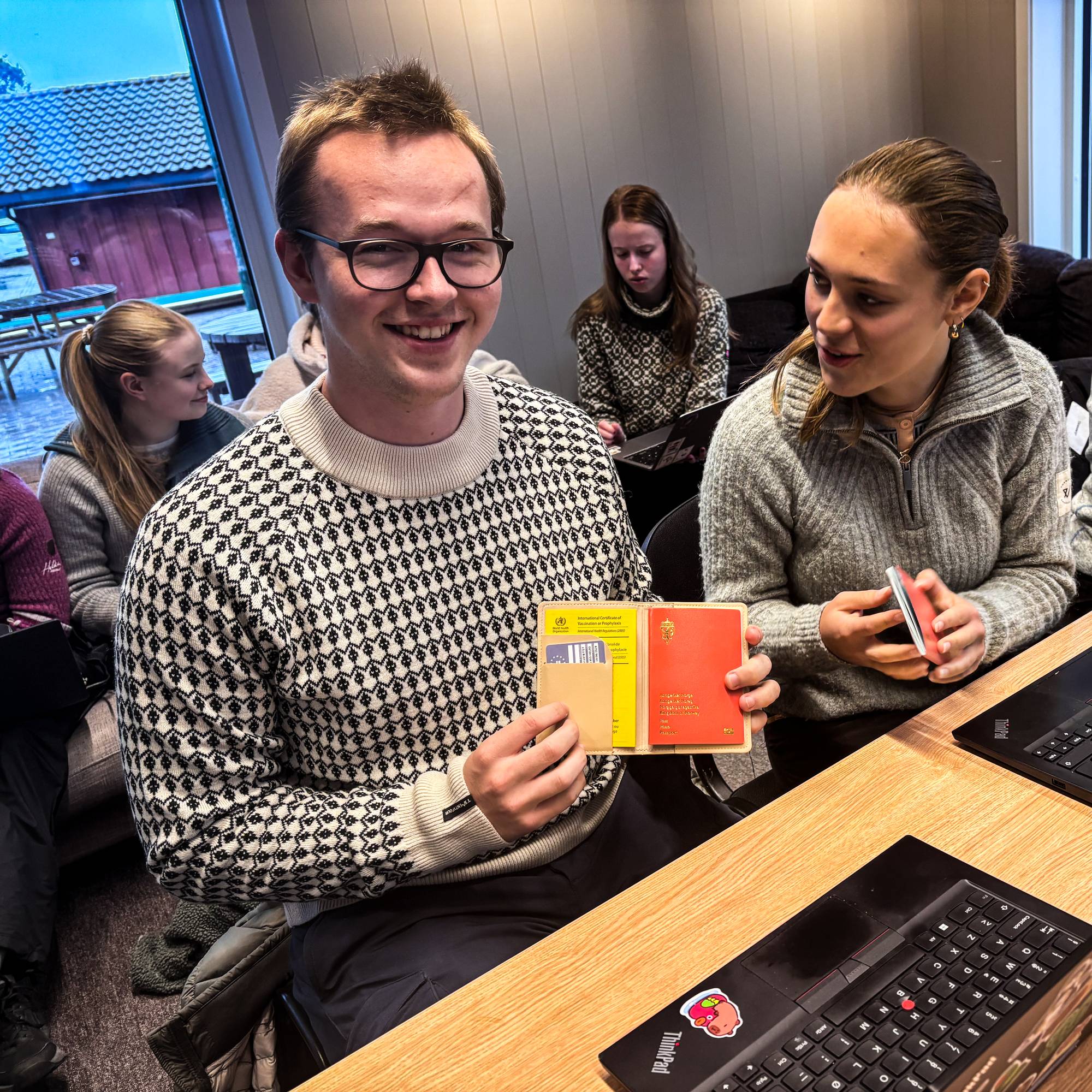 Studenter sitter ved et bord med laptoper. En mann viser frem et gult og rødt passetui. (Bildebeskrivelsen er laget av en KI-tjeneste)