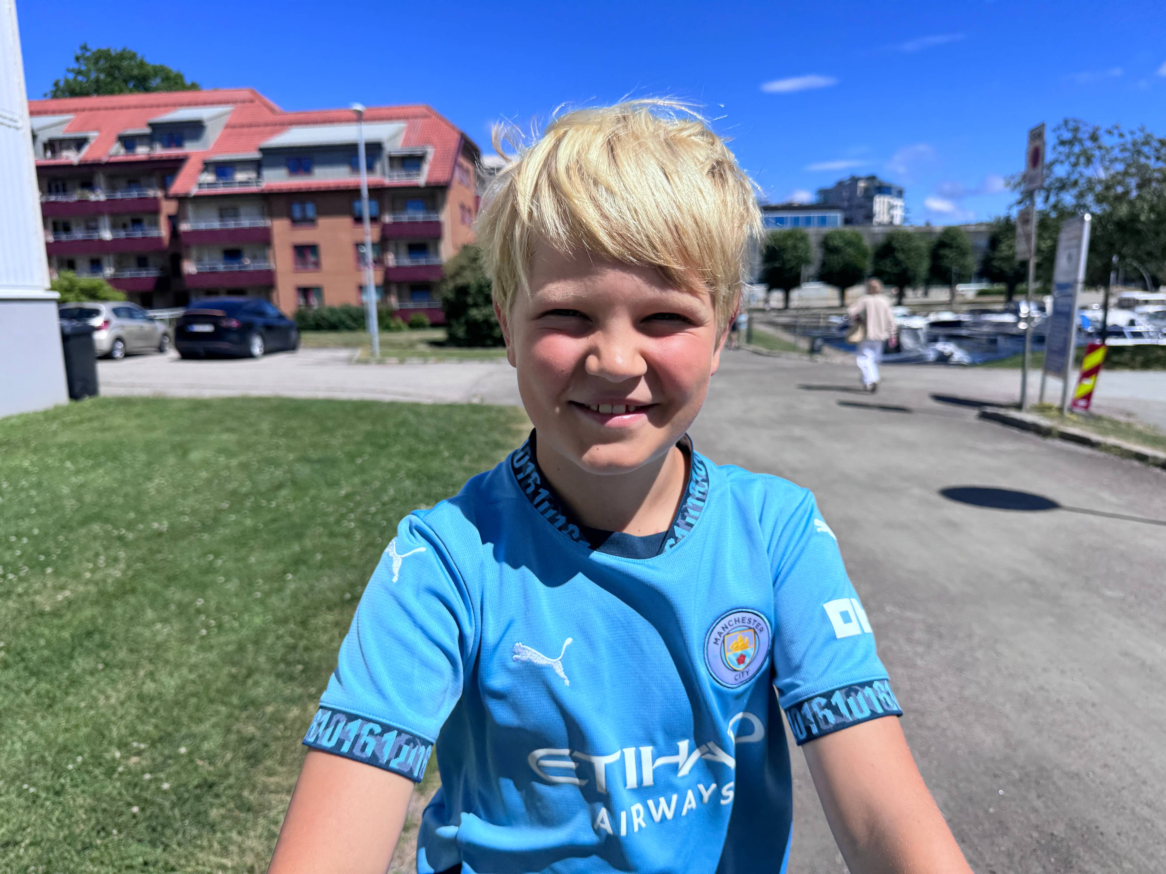 En gutt med blå fotballdrakt og lyst hår ser inn i kamera. I bakgrunnen ser man grønt gress og en grusvei.