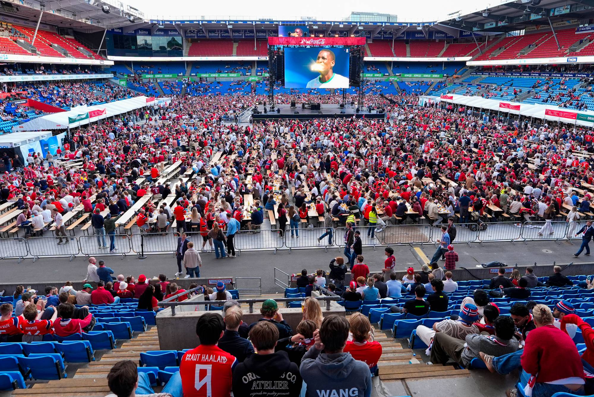 Stor folkemengde sitter på tribunen og ved langbord inne på en stadion. En stor skjerm viser et bilde av en mann. En scene er satt opp foran skjermen. (Bildebeskrivelsen er laget av en KI-tjeneste)