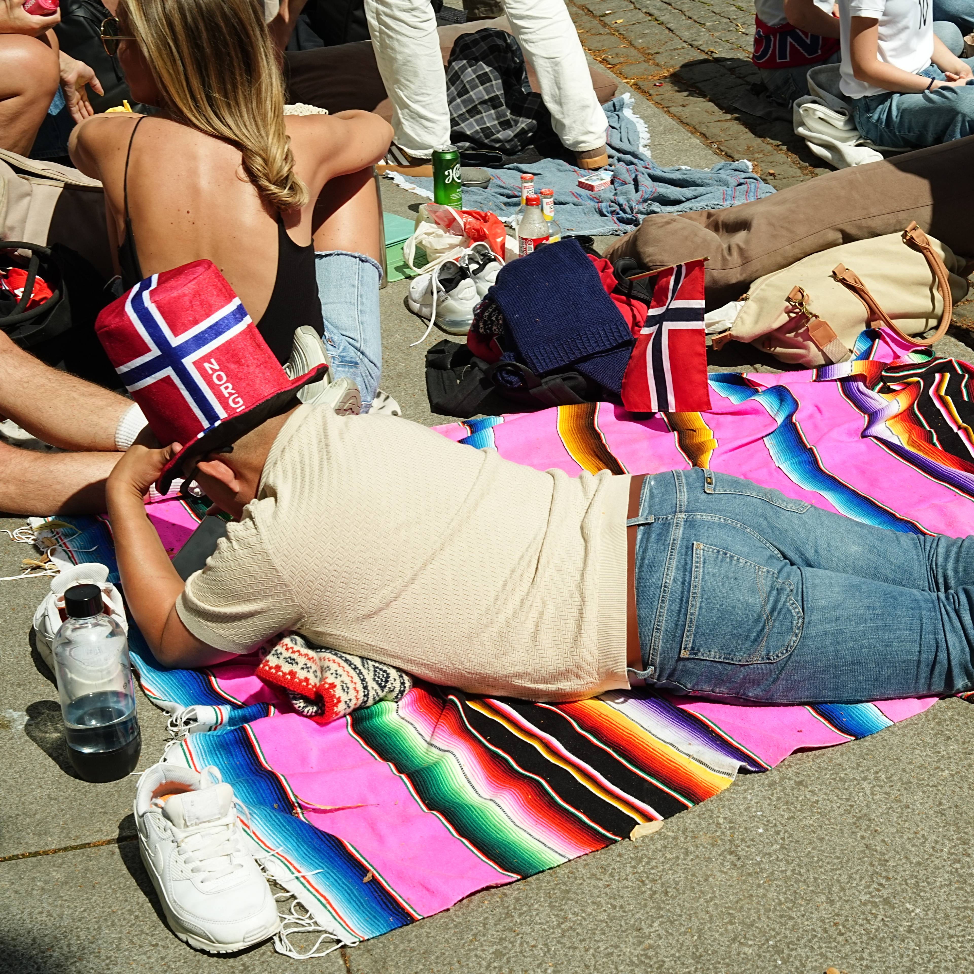 Person med norsk flagghatt ligger på et fargerikt teppe på bakken sammen med andre mennesker. (Bildebeskrivelsen er laget av en KI-tjeneste)