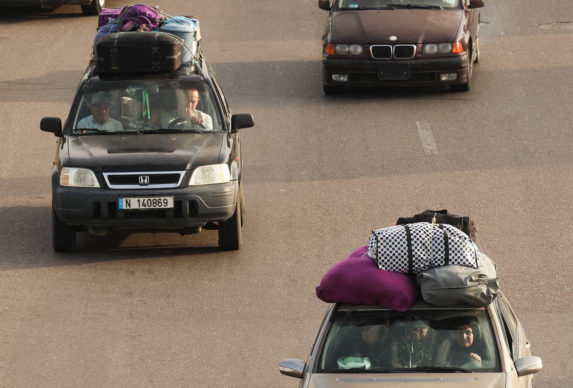 Biler med bagasje på taket kjører på en vei. To biler sees i bildet.
En svart bil kjører i forgrunnen. Den har flere kofferter og bager stablet på taket.
Inne i bilen sitter to personer. En bil med bagasje på taket kjører i bakgrunnen. (Bildebeskrivelsen er laget av en KI-tjeneste)