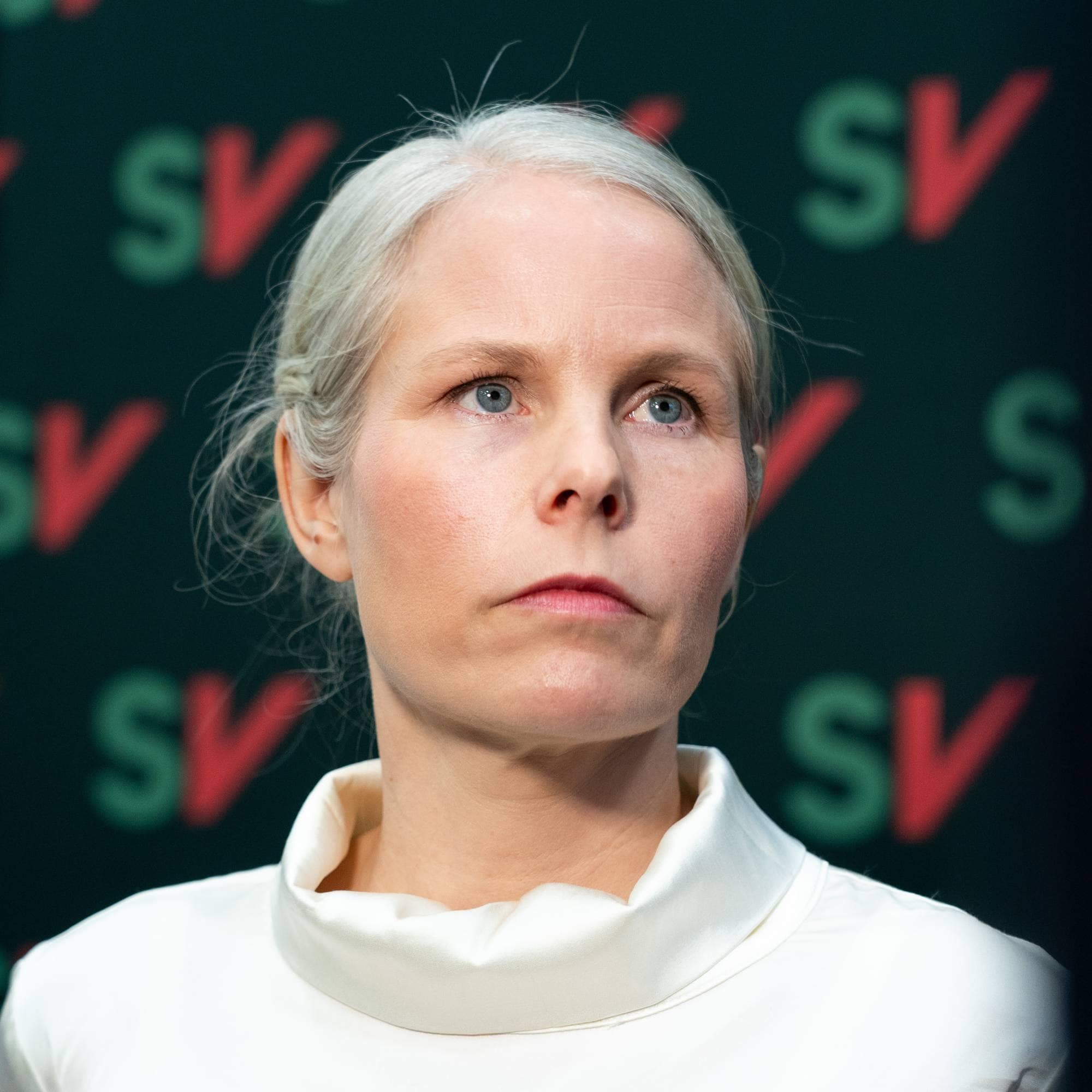 Kirsti Bergstø i hvit overdel ser alvorlig ut. I bakgrunnen en vegg med mange små SV-logoer på