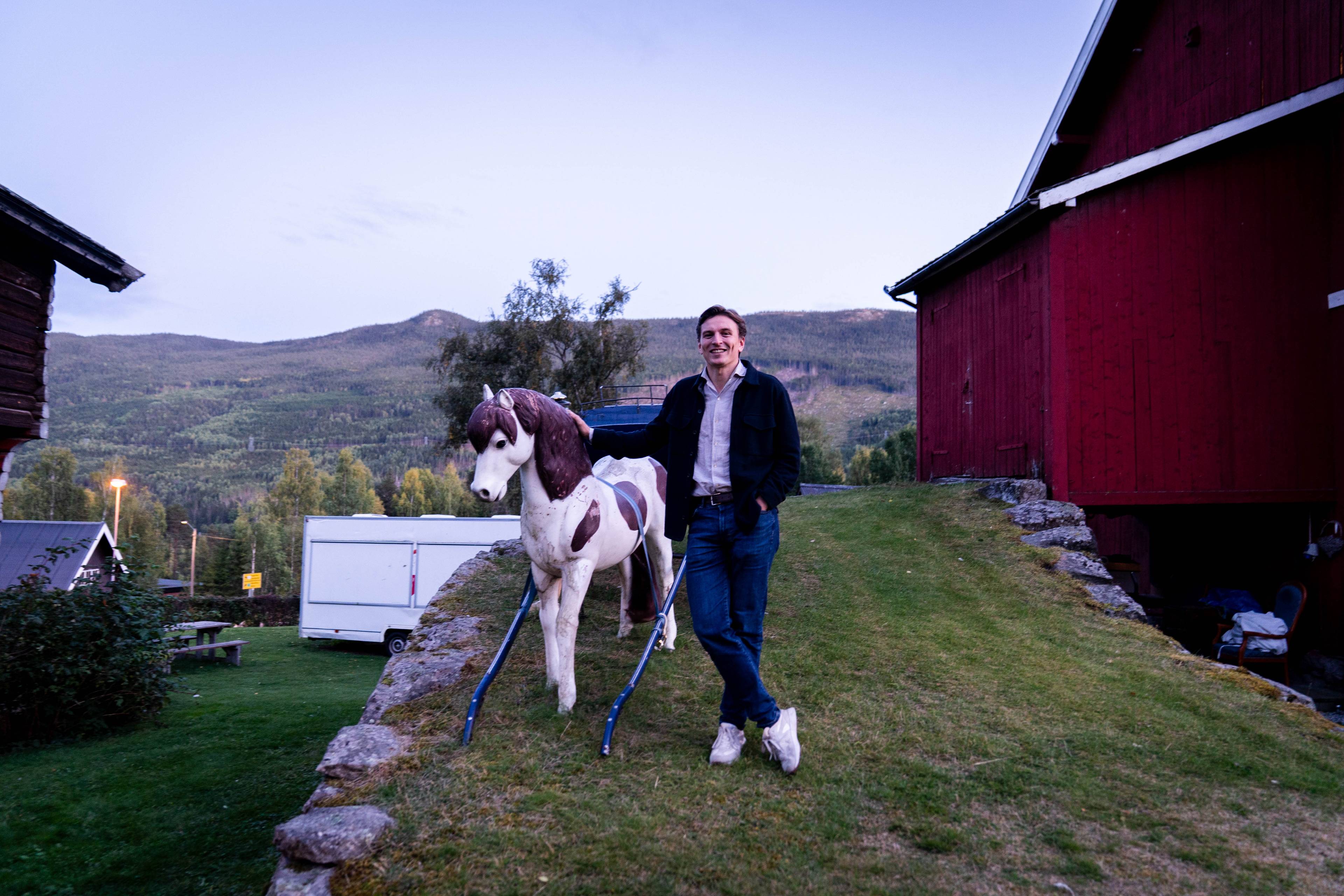 En mann står ved en lekehest på et gårdstun med røde låvebygninger i bakgrunnen. (Bildebeskrivelsen er laget av en KI-tjeneste)