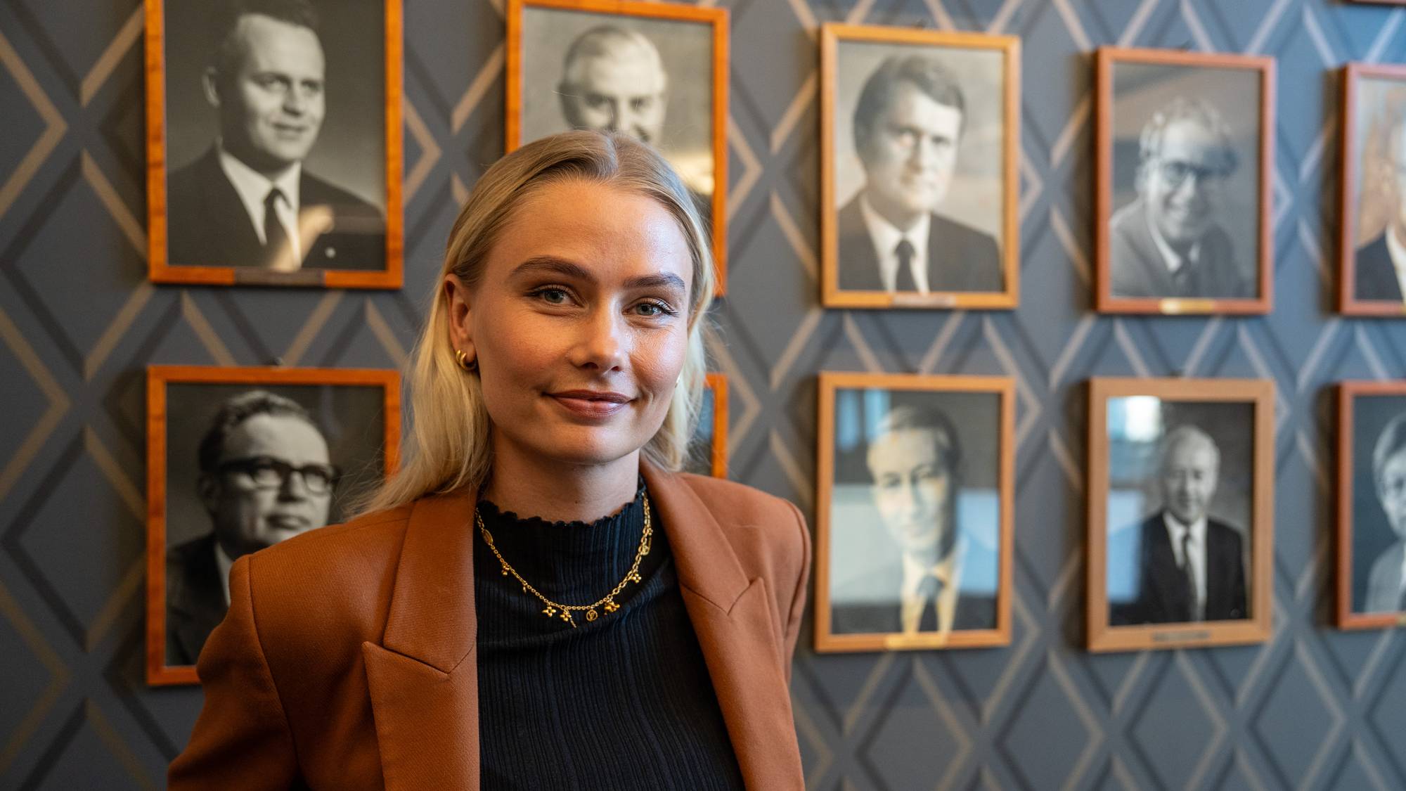 En kvinne står foran en vegg med innrammede portrettbilder. Hun er iført en brun blazer og et svart plagg. Bildet viser en samling av formelle portretter, trolig av tidligere ledere eller betydningsfulle personer. (Bildebeskrivelsen er laget av en KI-tjeneste)