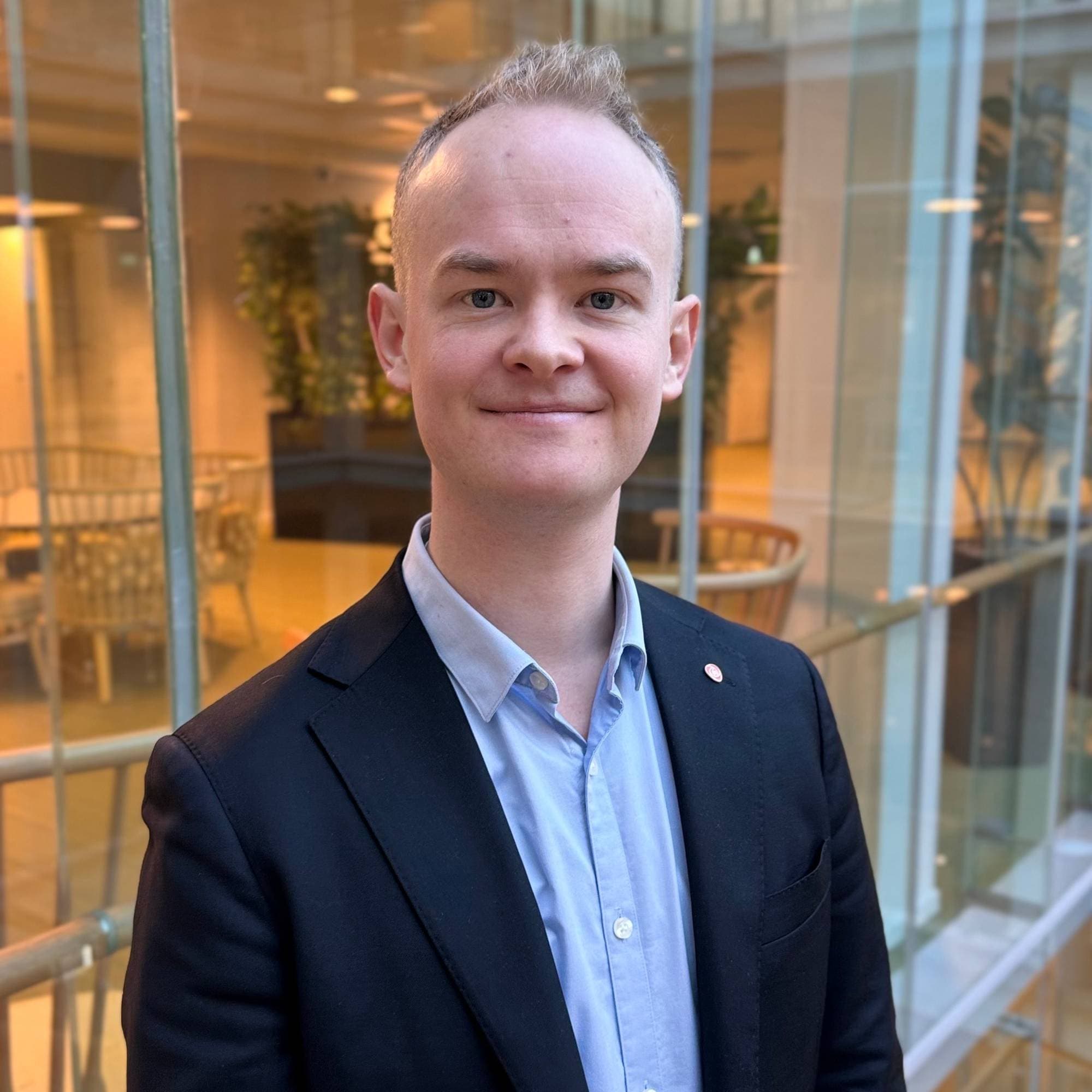 En mann i mørk dress og lyseblå skjorte står i en trapp. Han har et lite rødt merke på jakken. Bak ham er det glassvegger med en trapp og møbler synlig. (Bildebeskrivelsen er laget av en KI-tjeneste)