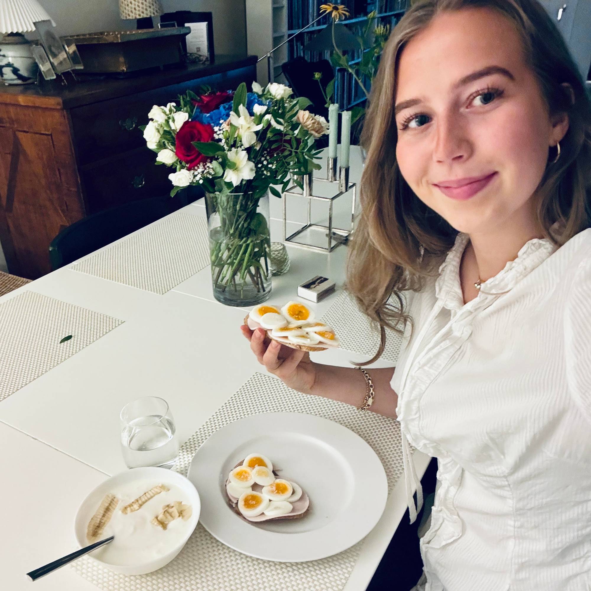Ung dame spiser frokost på et bord med hvit duk. På brødskiva har hun egg og skinke.