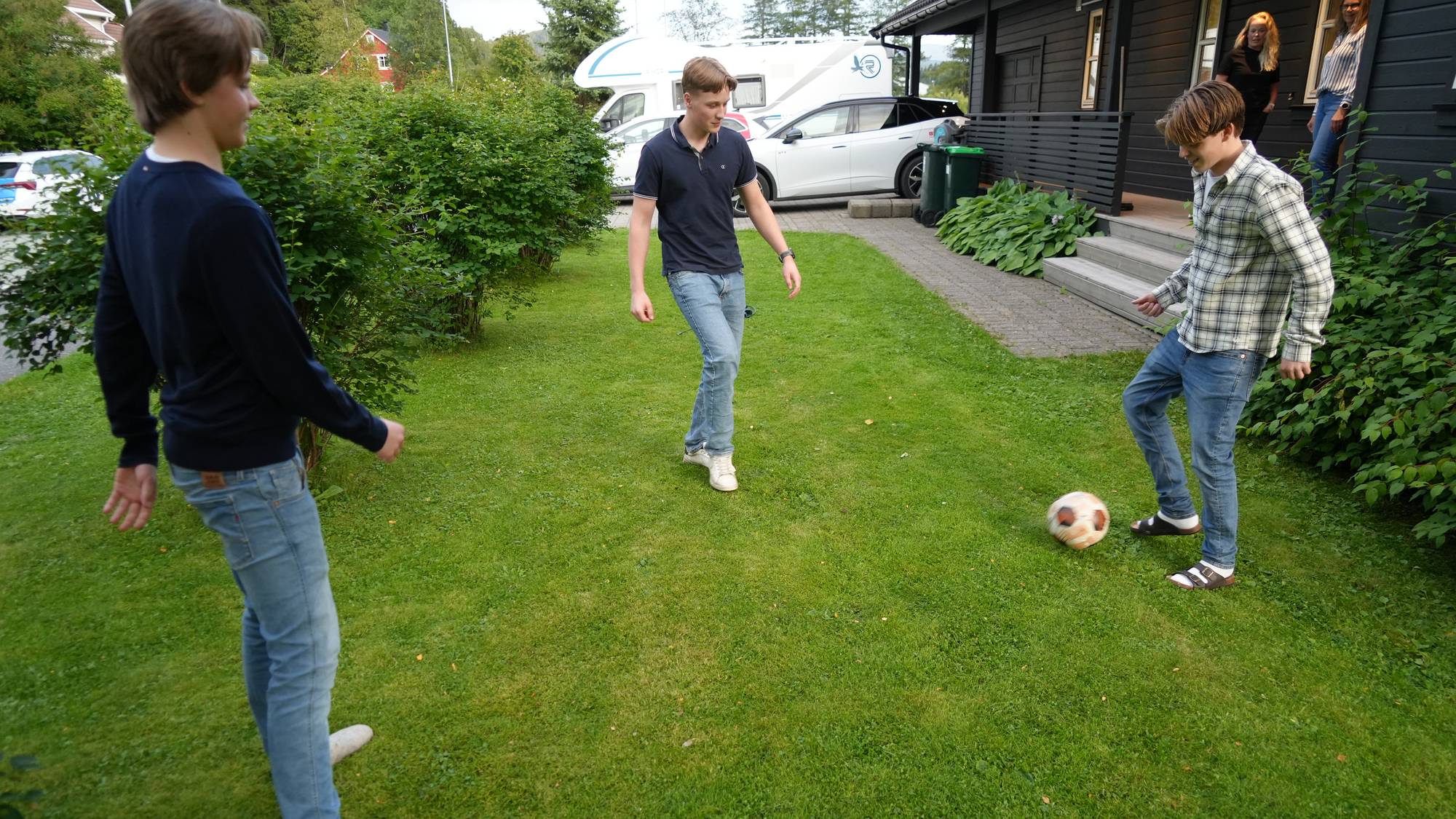Tre unge menn spiller fotball på en plen utenfor et hus. (Bildebeskrivelsen er laget av en KI-tjeneste)