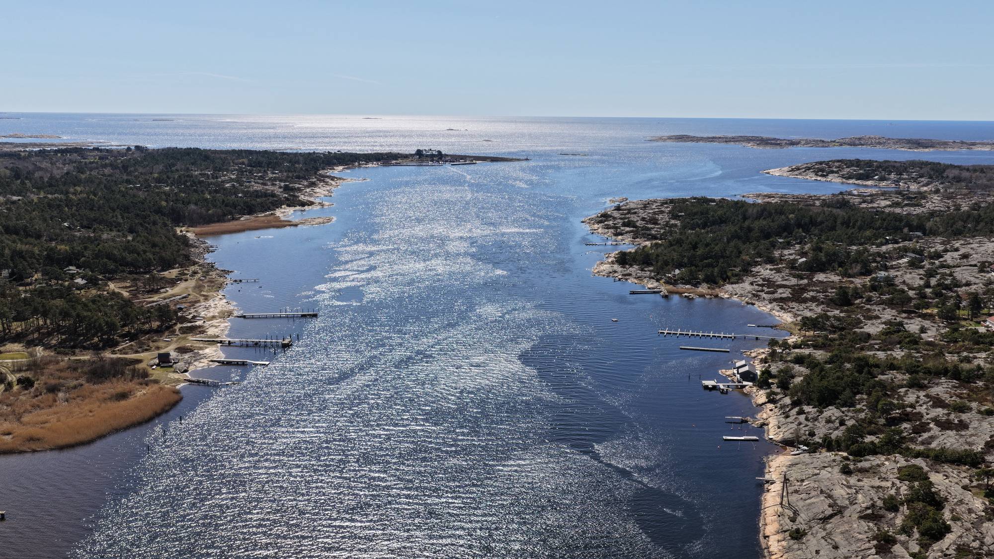 Luftfoto av en kyststripe med skogkledde øyer og holmer i solskinn. En bred vannvei, sannsynligvis en fjord eller bukt, strekker seg mot horisonten. Flere båter ligger fortøyd ved brygger langs den steinete kystlinjen. (Bildebeskrivelsen er laget av en KI-tjeneste)