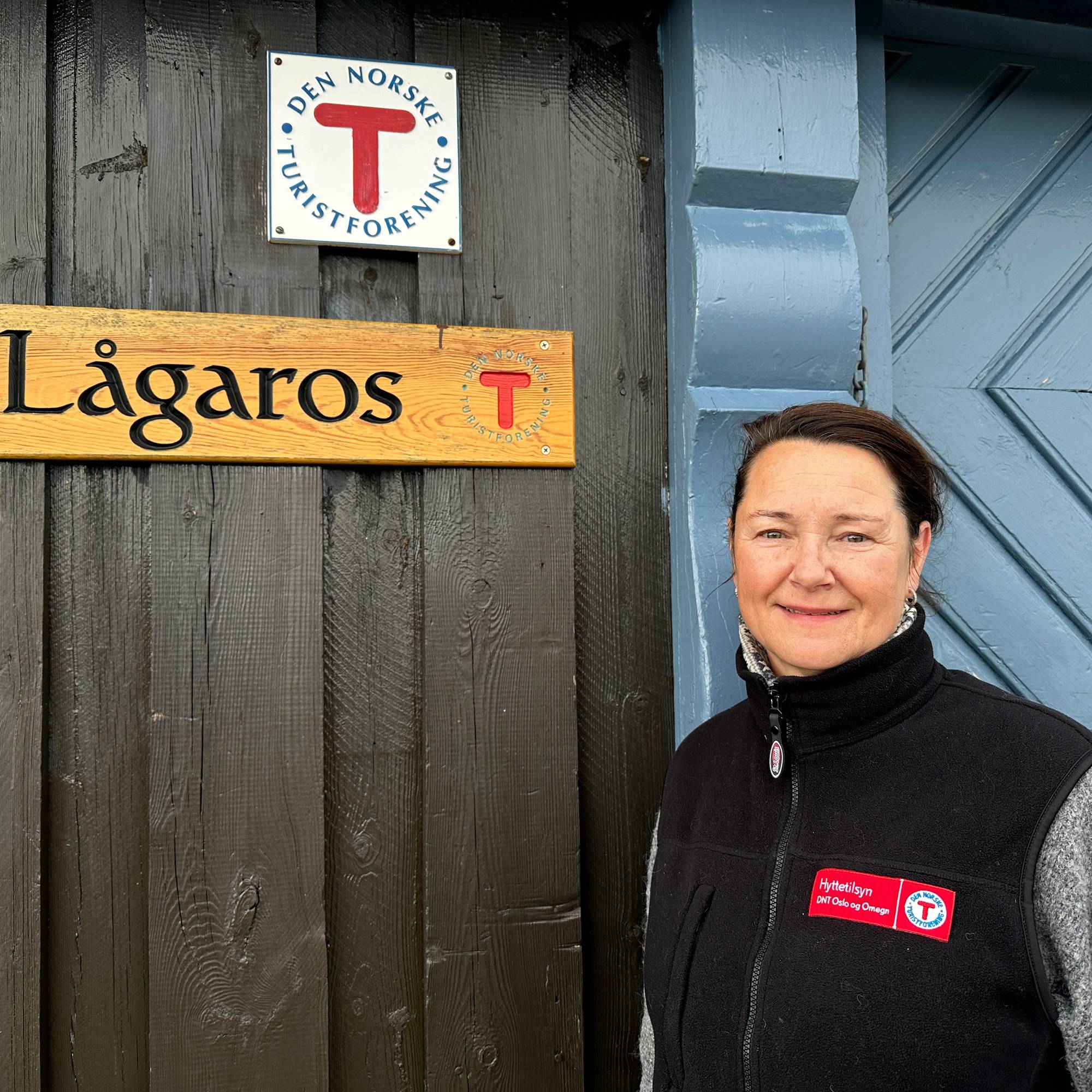 Kvinne med mørkt hår og DNT-vest foran skiltet til hytta Lågaros.