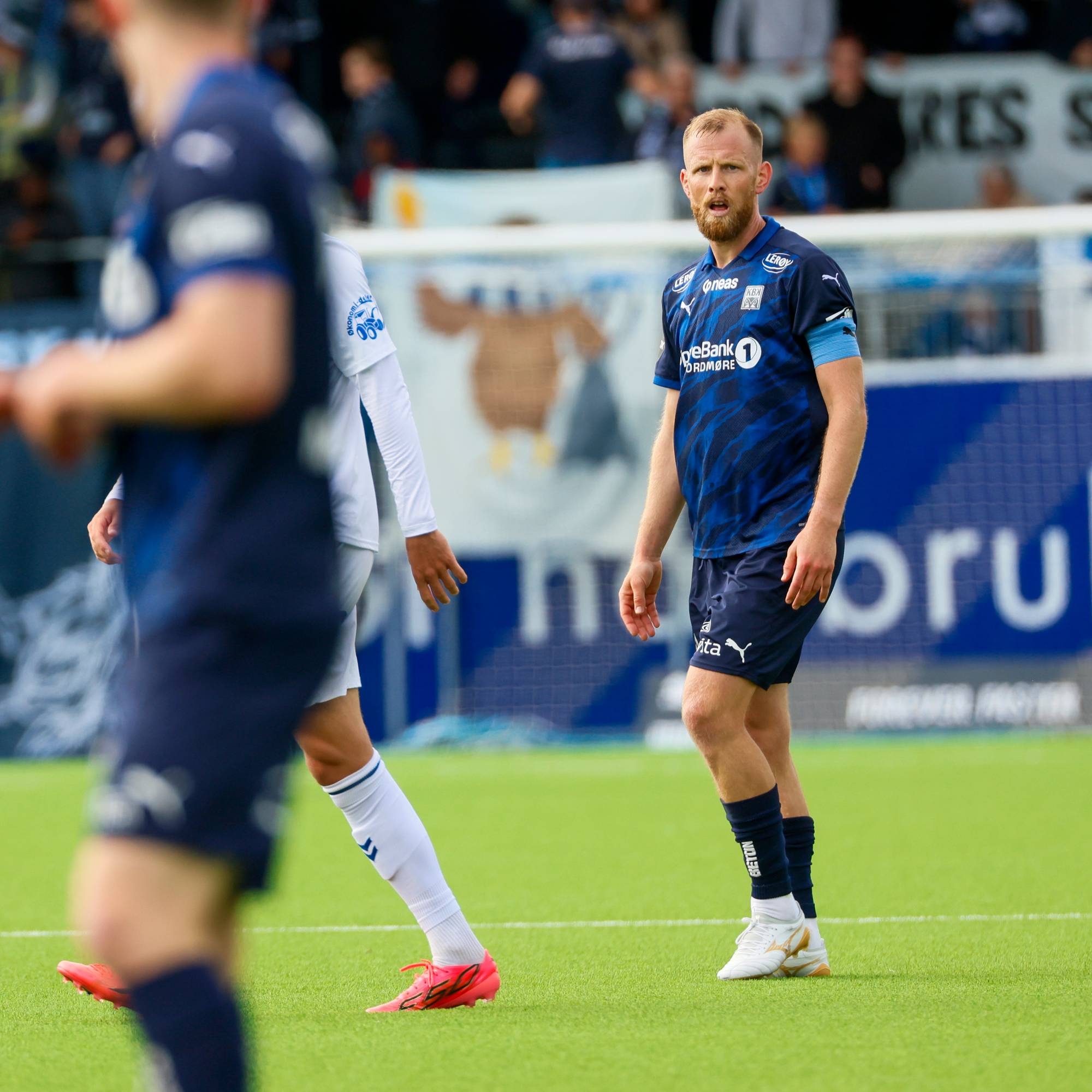 Fotballspillere på banen under en kamp. En spiller i blå drakt står foran tribunen. (Bildebeskrivelsen er laget av en KI-tjeneste)