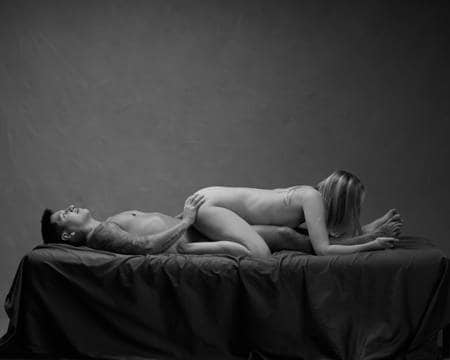 En naken mann med tatoveringer ligger på ryggen i senga og holder en naken dame på rumpa. Hun har lyst hår og ligger oppå ham med hode ned mot føttene hennes