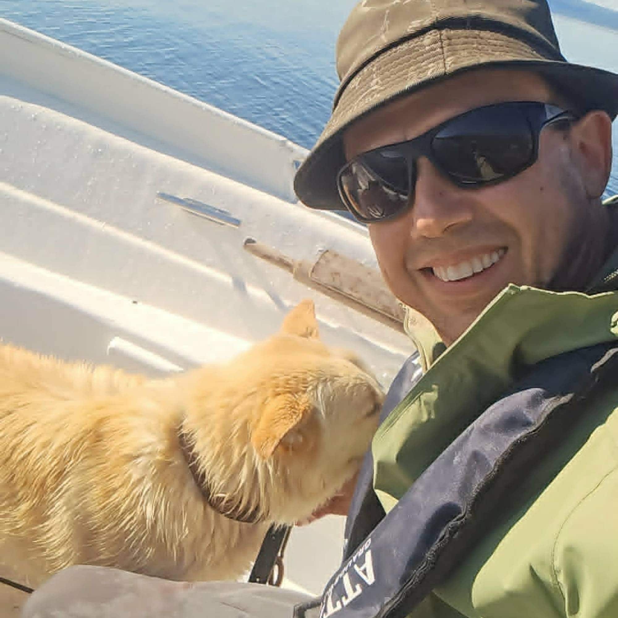 Person med solbriller og hatt sitter i en båt sammen med en hund på sjøen.