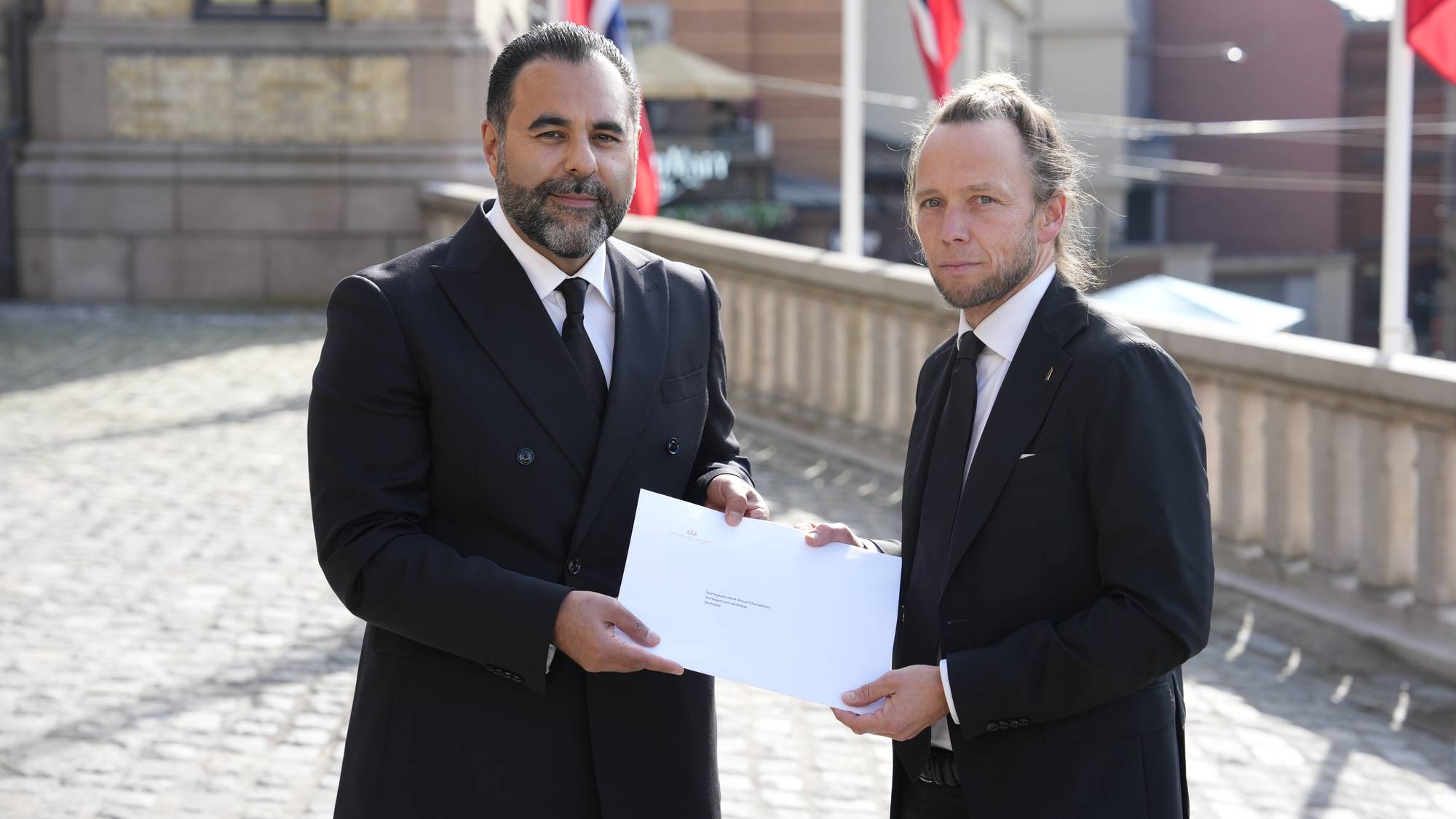 To menn i dress holder en konvolutt sammen foran Stortinget.