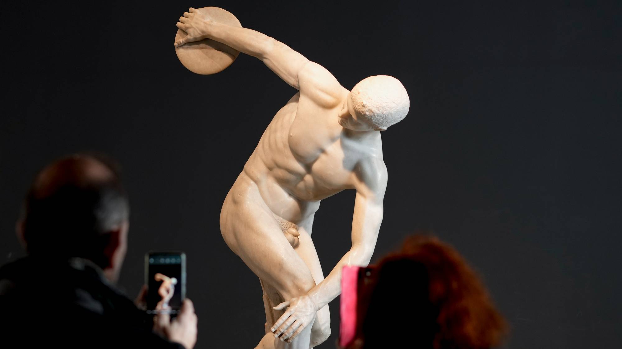 Museumsgjester fotograferer statuen Diskobolos mot en mørk bakgrunn. (Bildebeskrivelsen er laget av en KI-tjeneste)