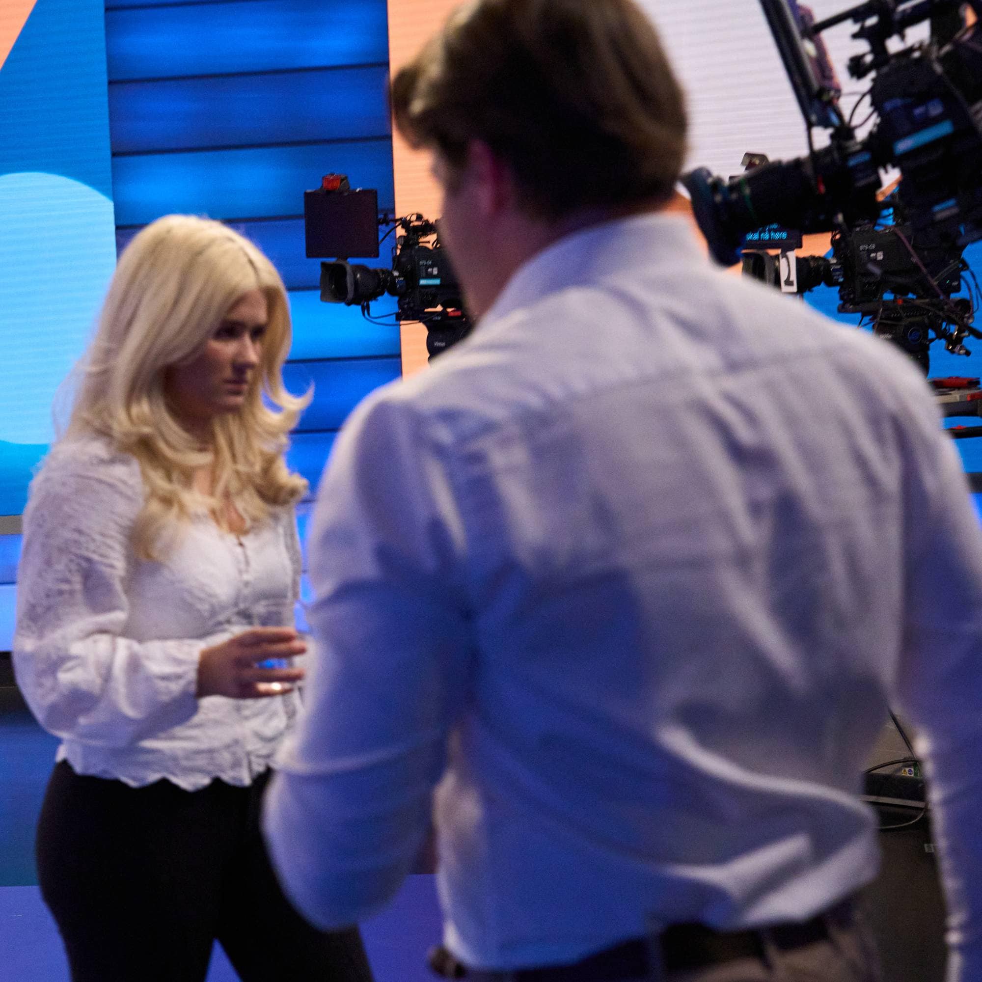 En kvinne og en mann står foran et TV-kamera i et studio med en fargerik grafisk skjerm i bakgrunnen. (Bildebeskrivelsen er laget av en KI-tjeneste)