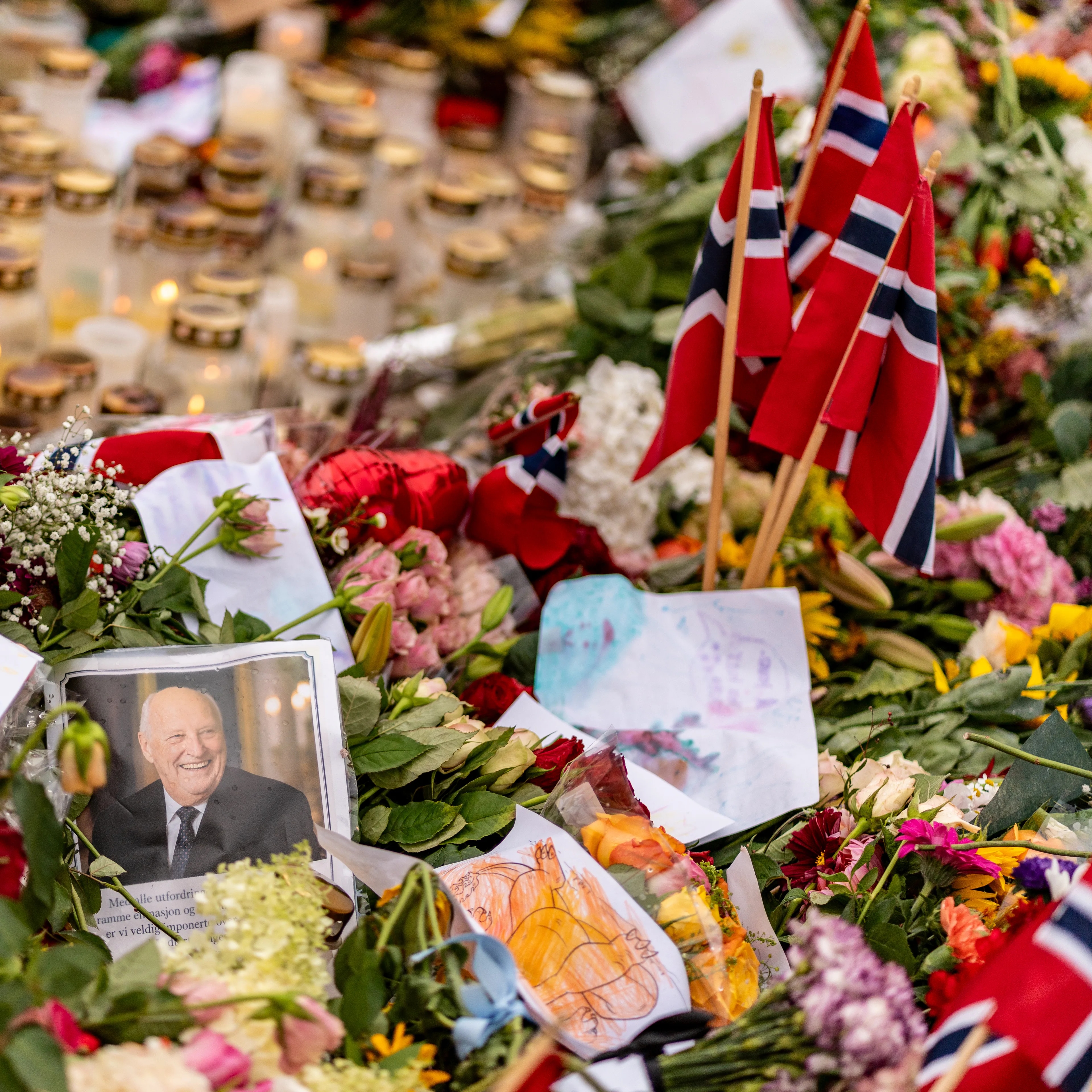 Blomster, tente lys og små norske flagg lagt ned foran et portrettbilde av Kong Harald. (Bildebeskrivelsen er laget av en KI-tjeneste)