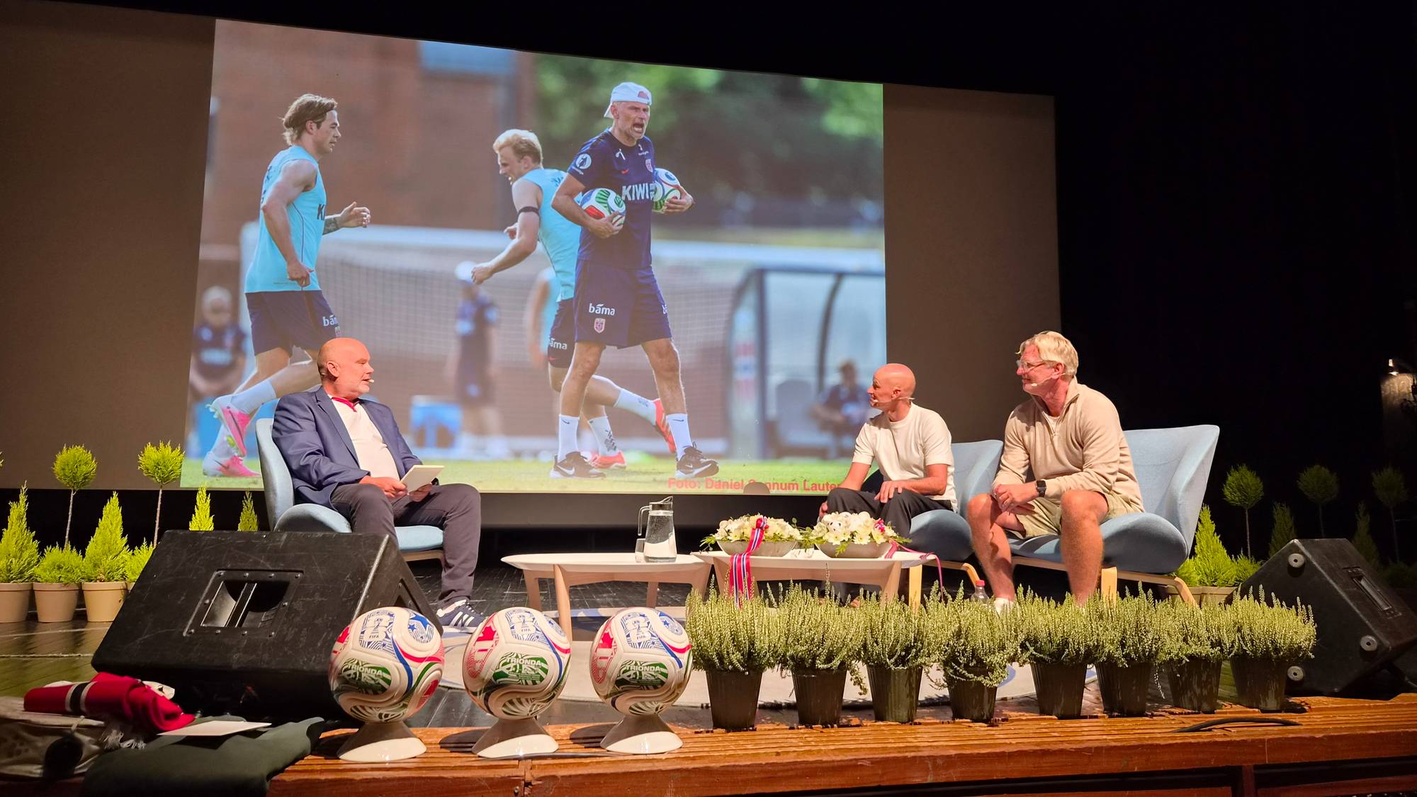 Tre menn sitter i stoler på en scene under en samtale, med et stort bilde av fotballtrening bak. (Bildebeskrivelsen er laget av en KI-tjeneste)
