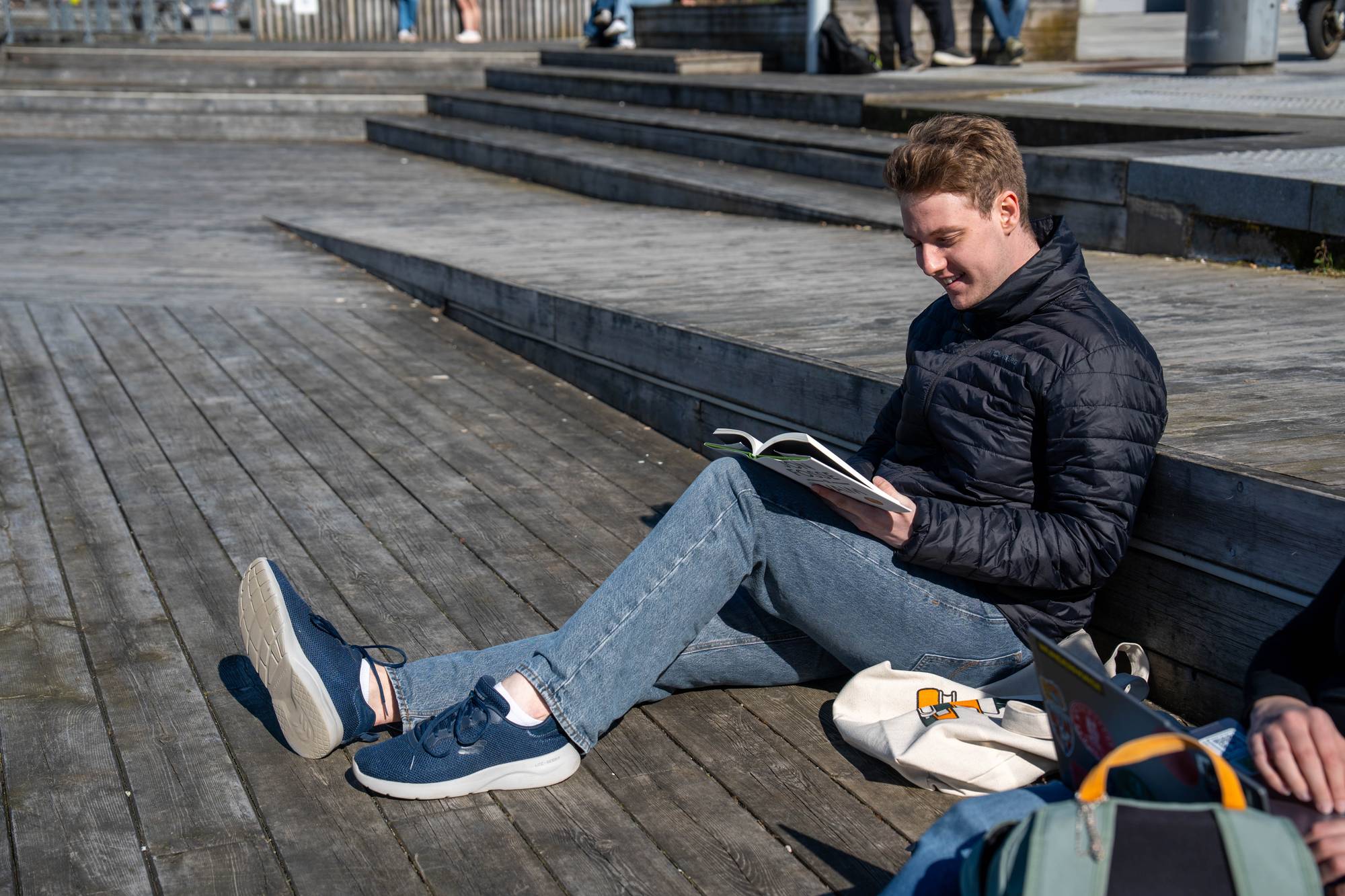En ung mann sitter på et tredekk og leser en bok. Han har på seg en svart dunjakke og blå jeans. I bakgrunnen er det trapper og flere personer. En hvit bag med tegninger ligger ved siden av mannen. (Bildebeskrivelsen er laget av en KI-tjeneste)