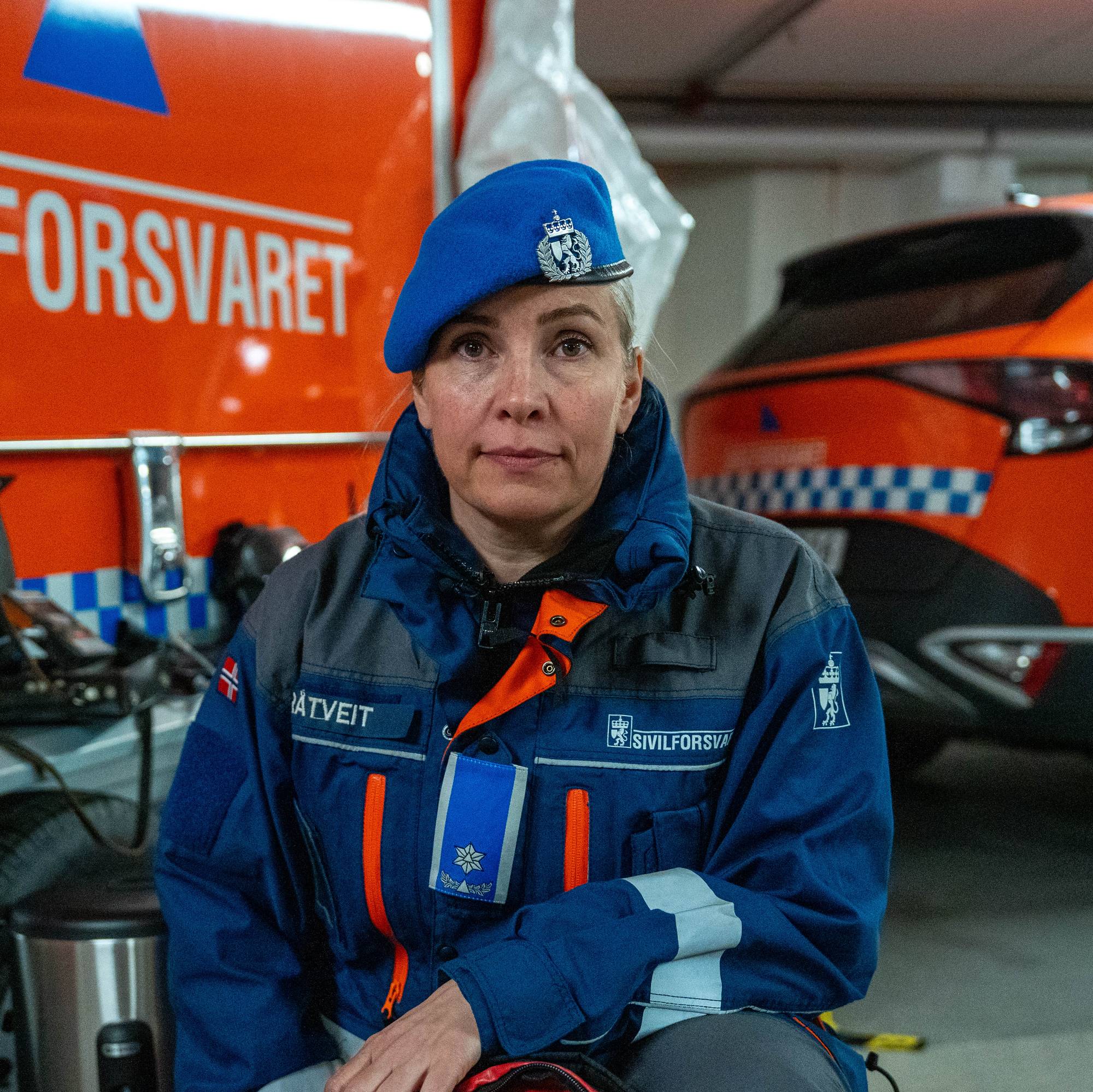 En kvinne med uniform sitter foran en oransje bil fra Sivilforsvaret. Hun har på seg en blå beret.