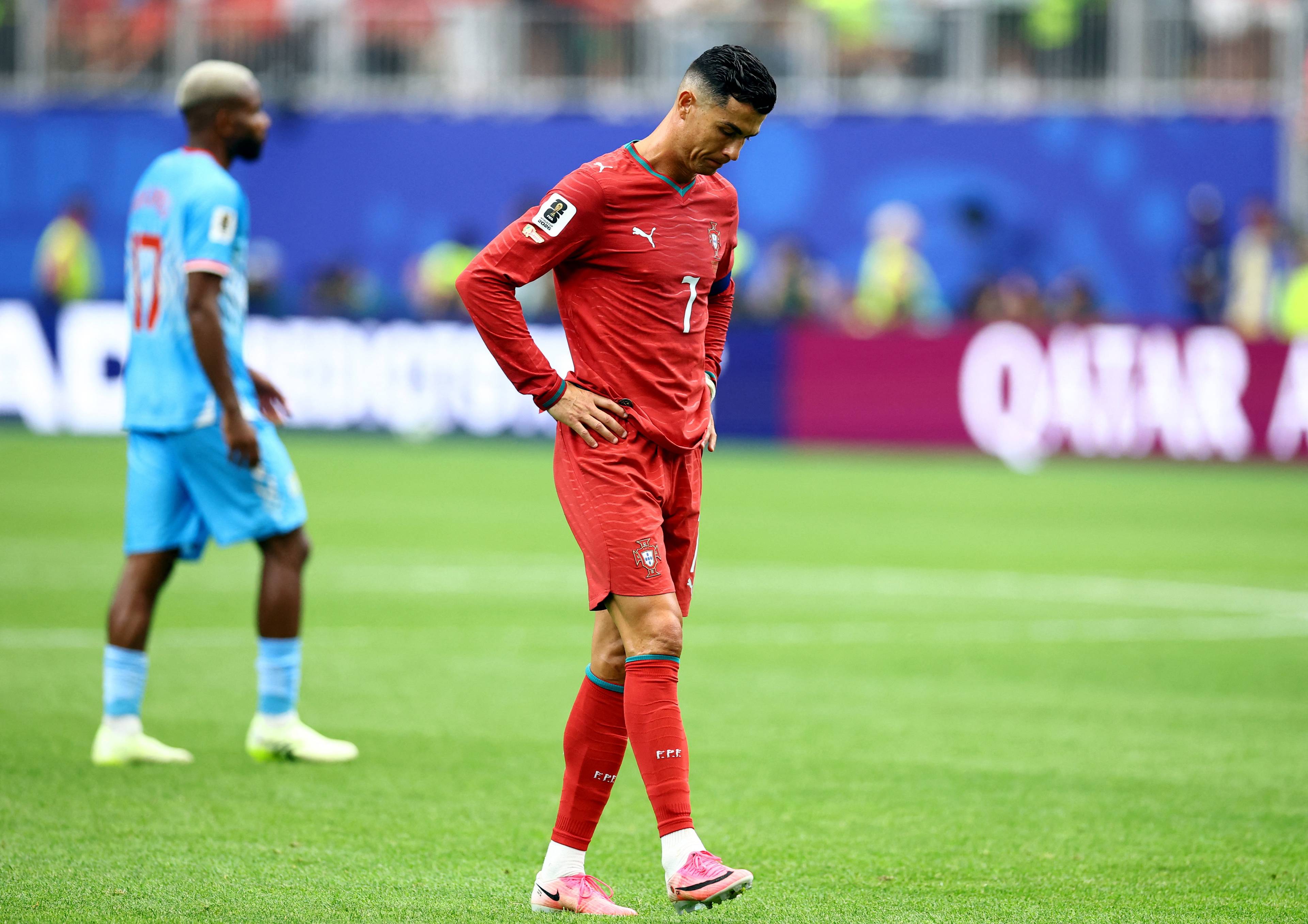 Fotballspiller Cristiano Ronaldo i rød drakt står med hendene på hoftene. En annen spiller i blå drakt står til venstre. Bakgrunnen viser tribuner og et QATAR AIRWAYS-banner. (Bildebeskrivelsen er laget av en KI-tjeneste)